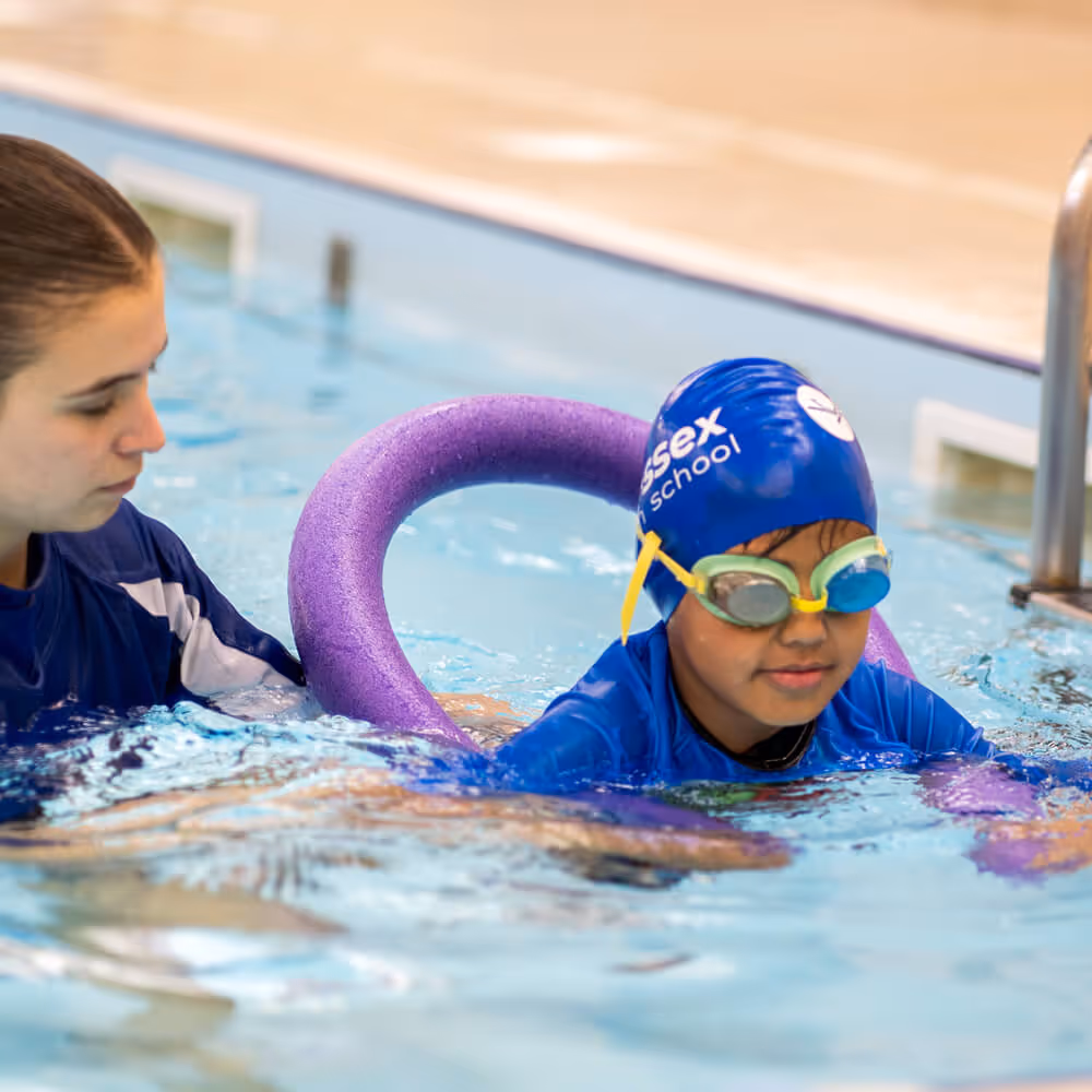 A London Swim School swim instructor assists a child wearing a blue swim cap and goggles using a purple pool noodle for support in a swimming pool.