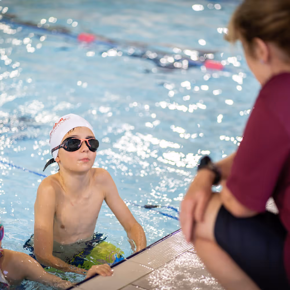 Young boy in a swim cap and goggles listening to a coach at the edge of a swimming pool.