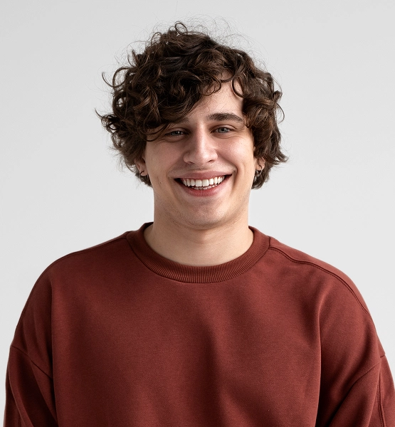 Smiling young man with curly dark hair wearing a rust-colored sweatshirt against a plain light background.