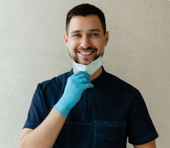 Smiling man with short dark hair and beard wearing blue medical scrubs and gloves, pulling down a surgical mask.