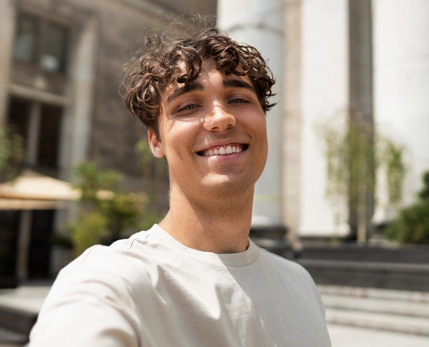 Young man with curly hair smiling outdoors in front of steps and a building.