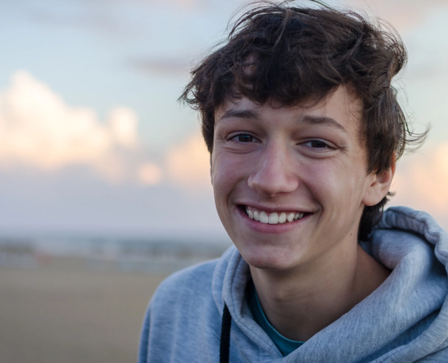 Smiling teenage boy with tousled hair wearing a gray hoodie, with a blurred outdoor background at sunset.