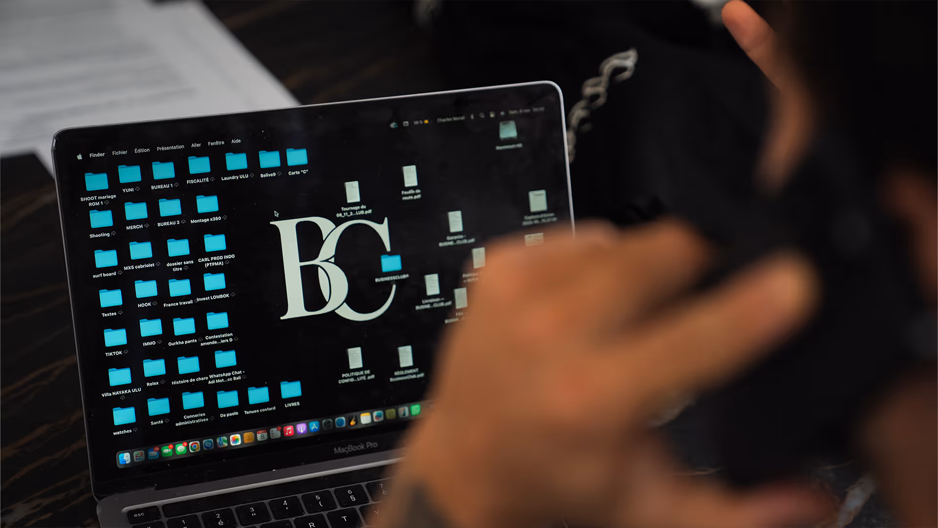 Personne tenant un téléphone portable en face d'un MacBook Pro affichant un bureau avec des dossiers et des fichiers.