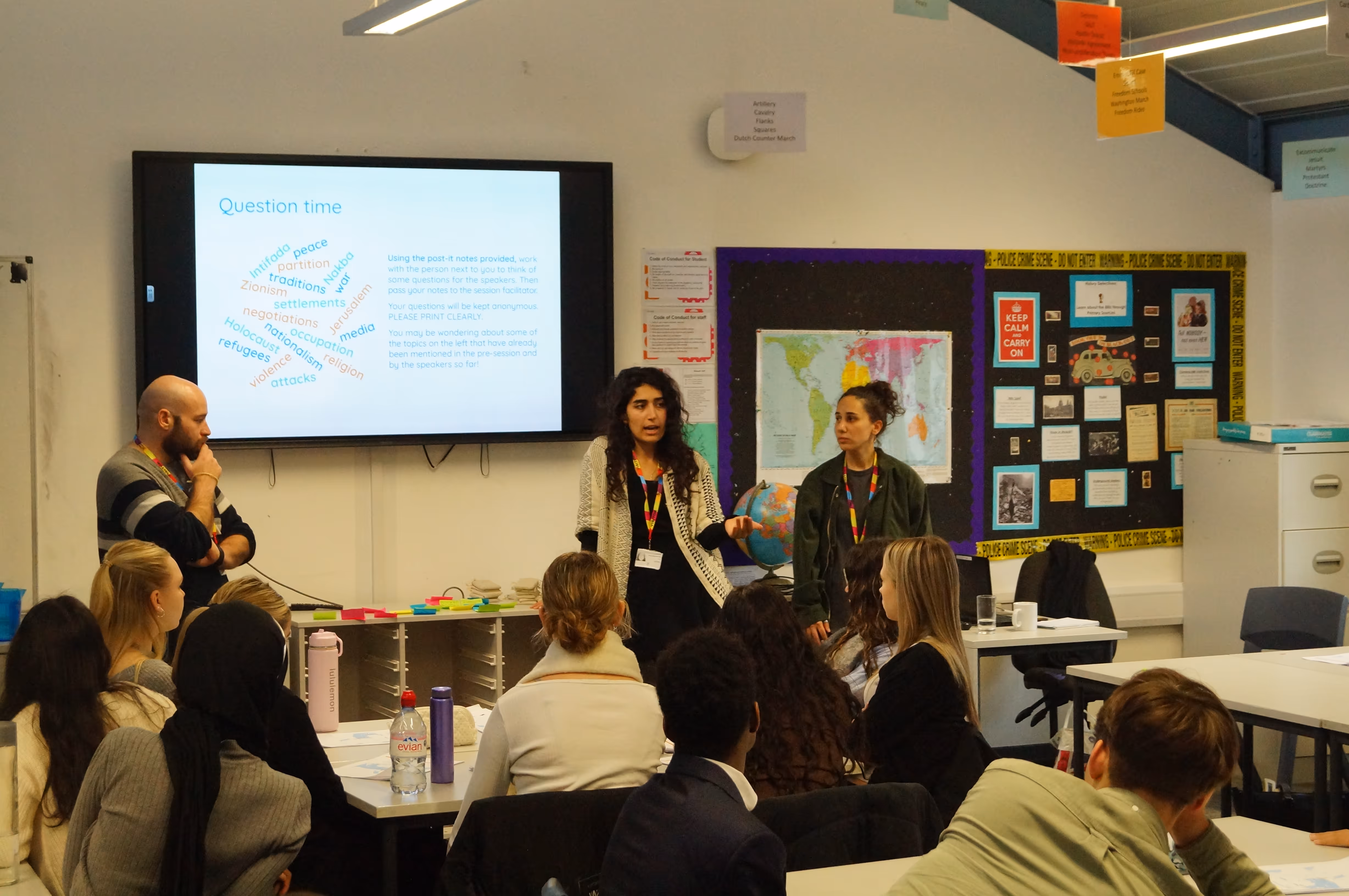 Classroom scene with a male and two female facilitators leading a discussion in front of a screen showing keywords about peace and conflict, while students listen seated around tables.