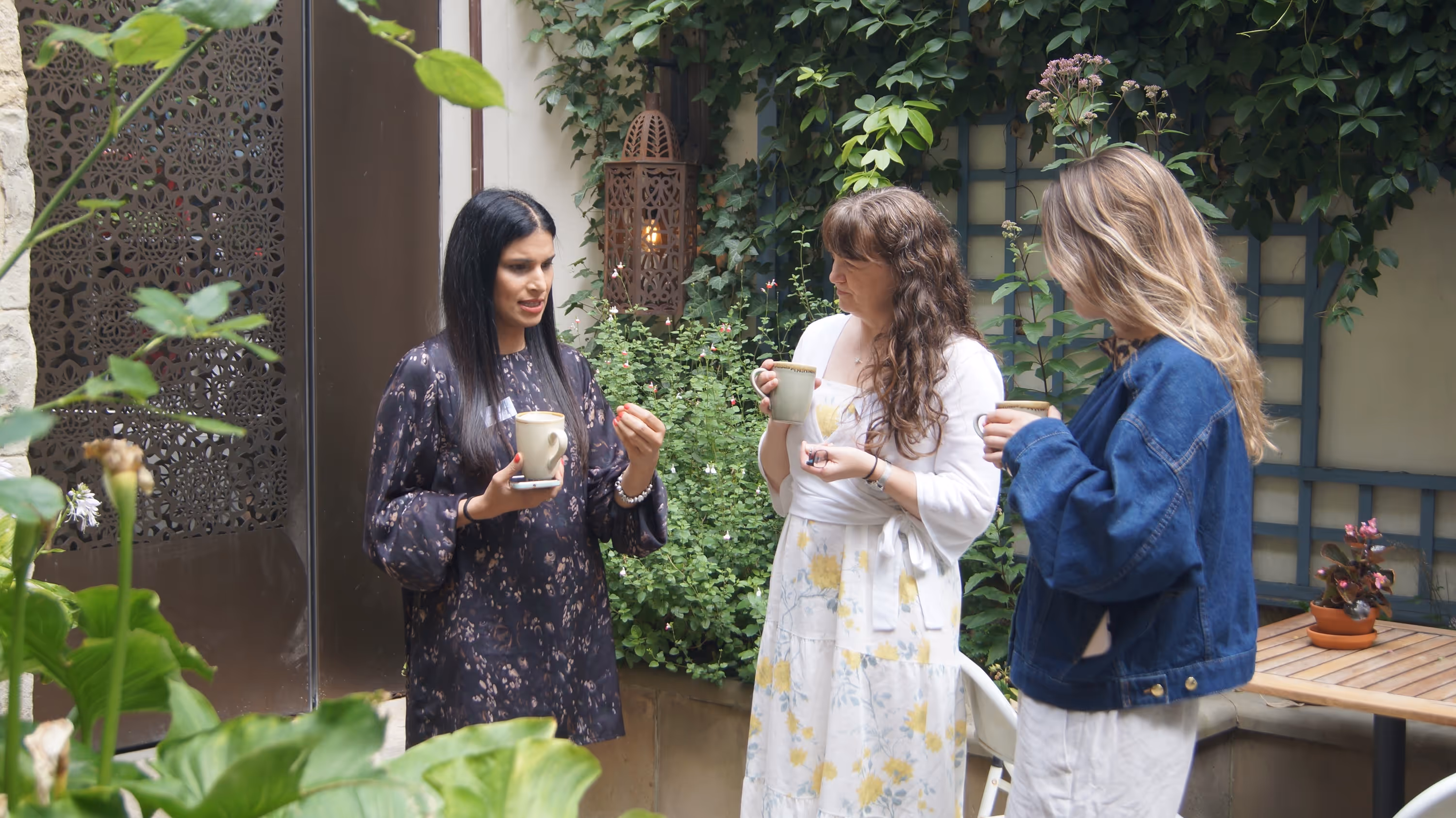Three women holding mugs and talking in a garden with green plants and a wooden table.