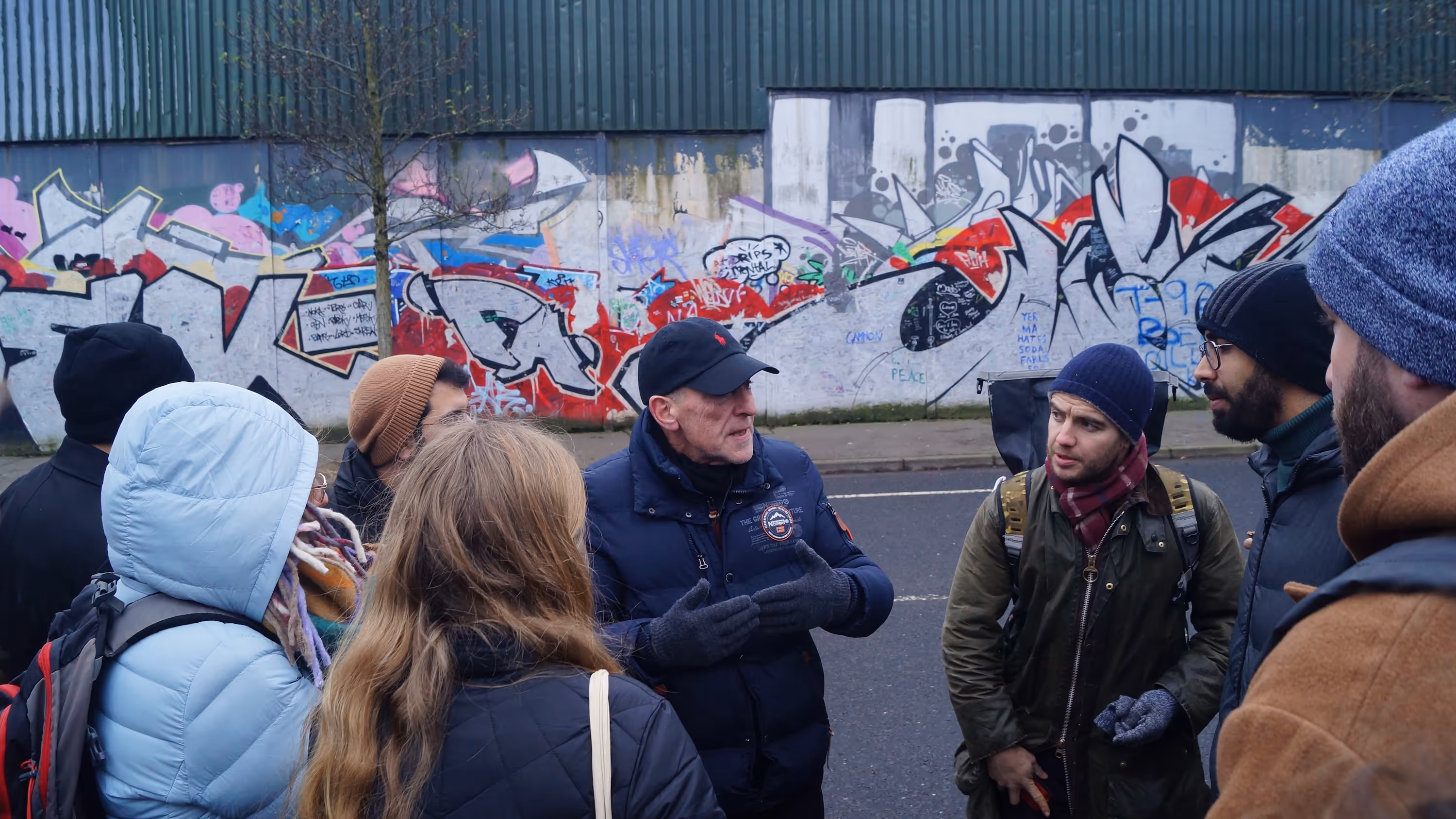 A group of people dressed in winter clothing listening to an older man speaking on a street with a graffiti-covered wall in the background.