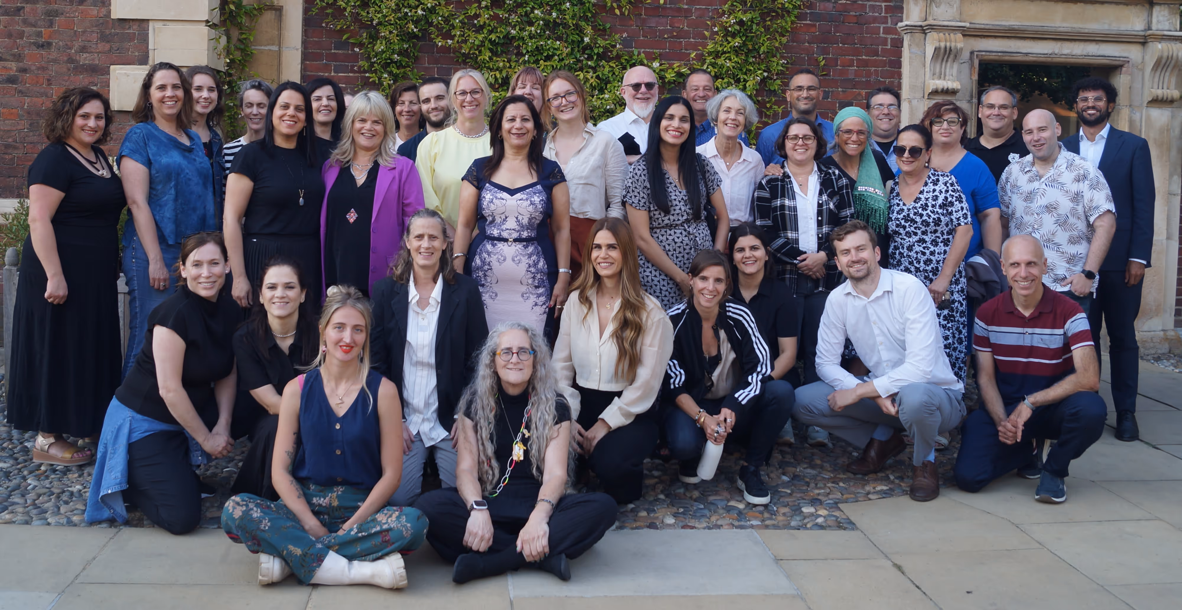 Group of smiling adults posing outdoors in front of a brick wall with greenery.
