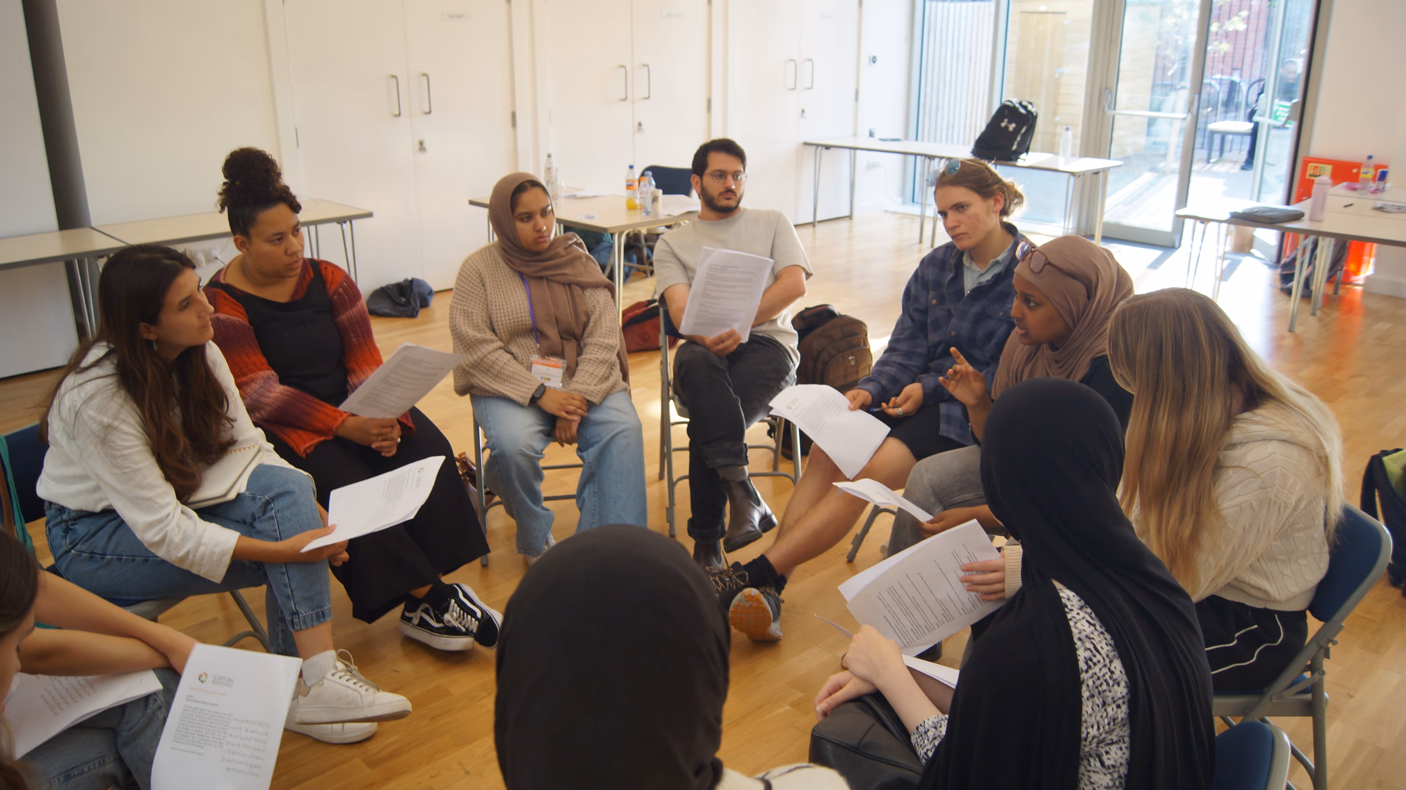 Diverse group of people sitting in a circle in a bright room, holding and discussing papers.