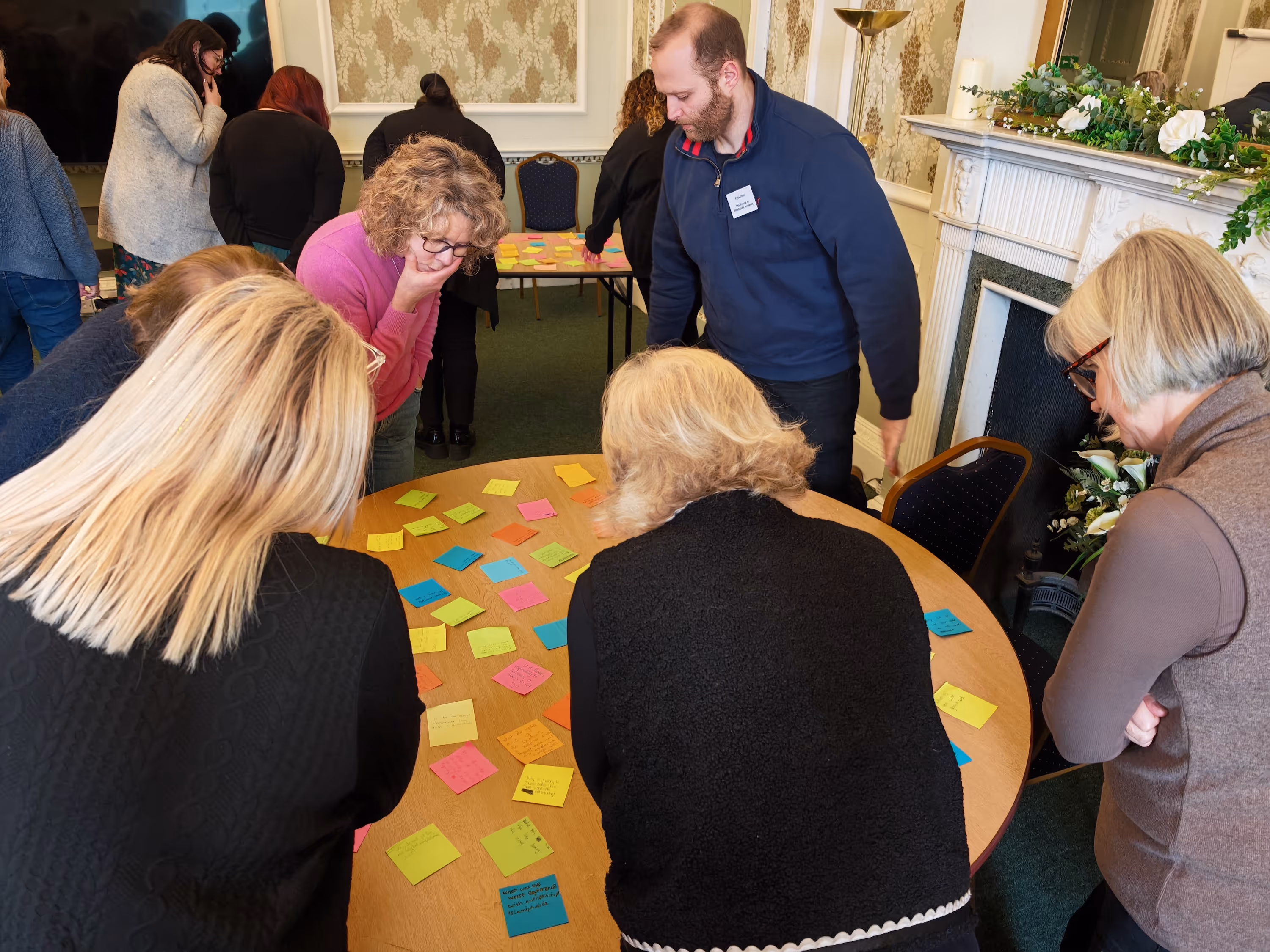 Group of people gathered around a round table covered with colorful sticky notes in a meeting room with ornate wallpaper and a fireplace.