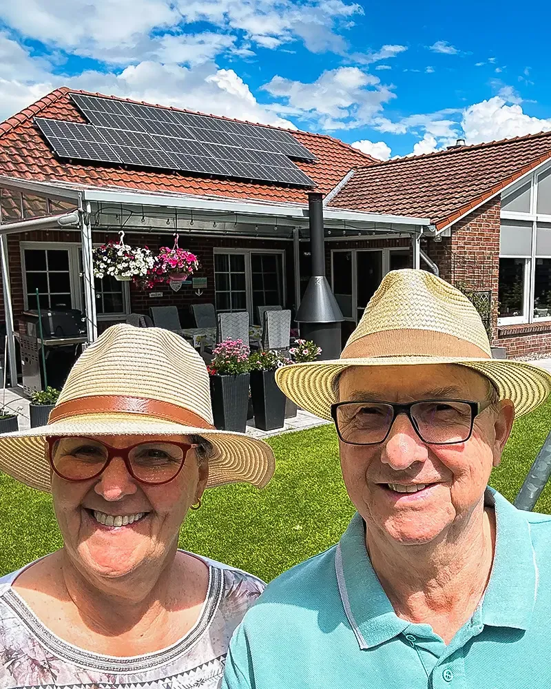 Älteres Paar mit Strohhüten und Brillen lächelt vor einem Haus mit Solarpaneelen auf dem Dach und einem blühenden Garten.