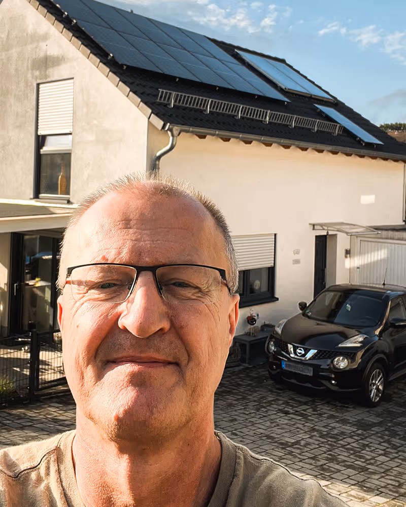 Mann mit Brille vor einem Haus mit Solarpanelen auf dem Dach und einem schwarzen Auto auf dem Hof.