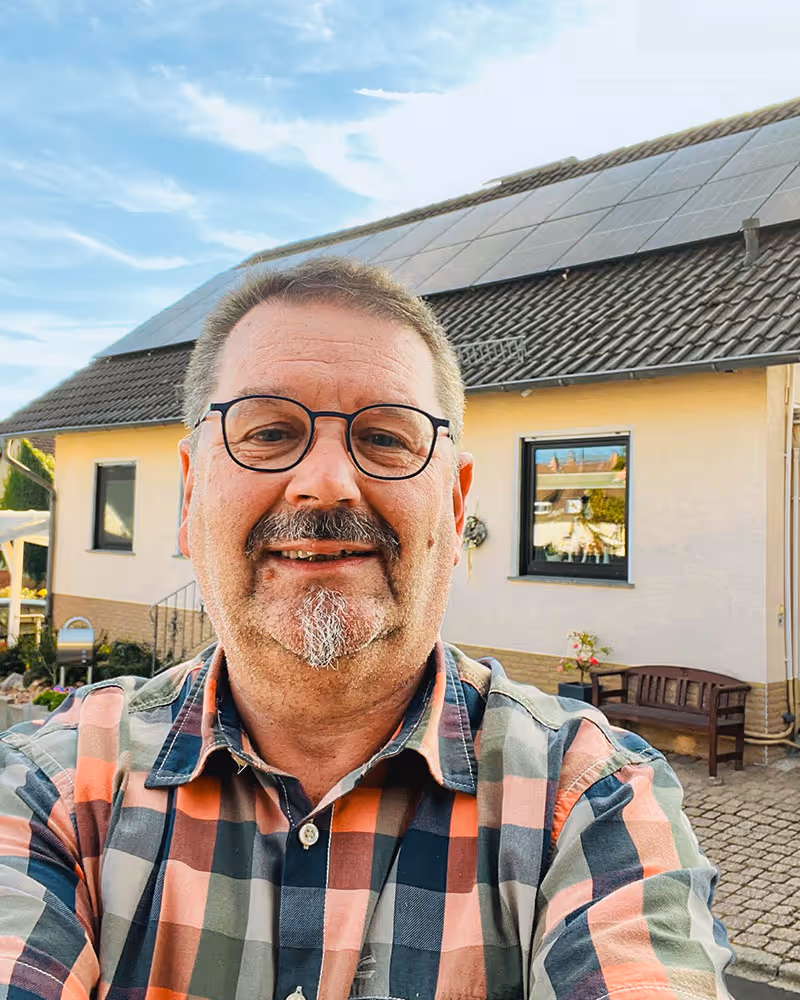 Ein Mann mit Brille und kariertem Hemd macht ein Selfie vor einem Haus mit Solarpaneelen auf dem Dach.