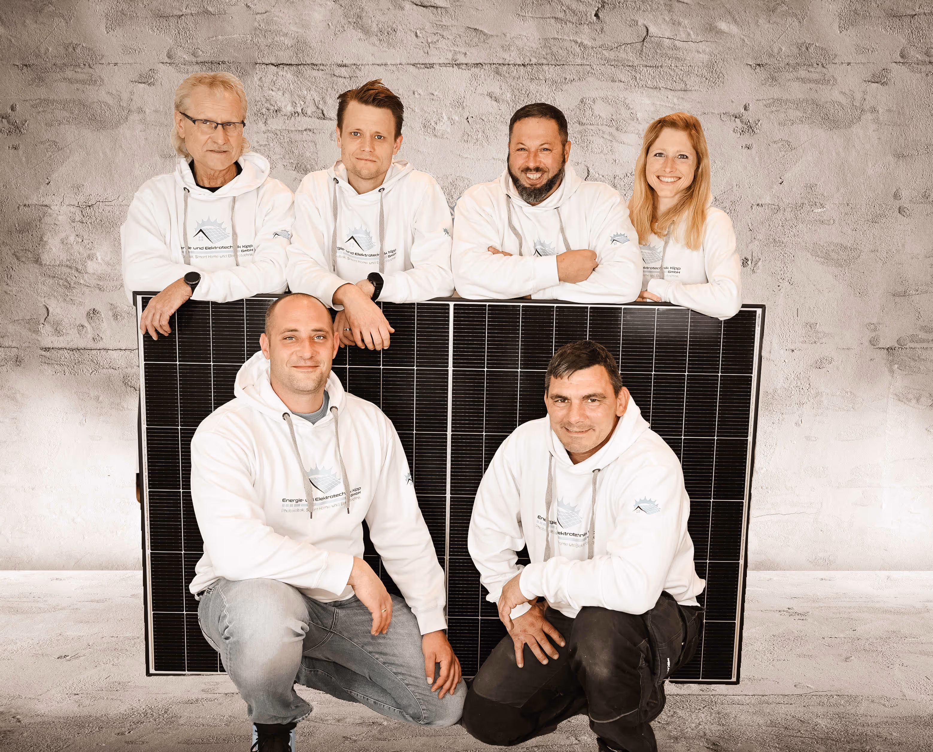 Sechs Personen in weißen Hoodies posieren mit großem Solarmodul vor grauer Mauer.