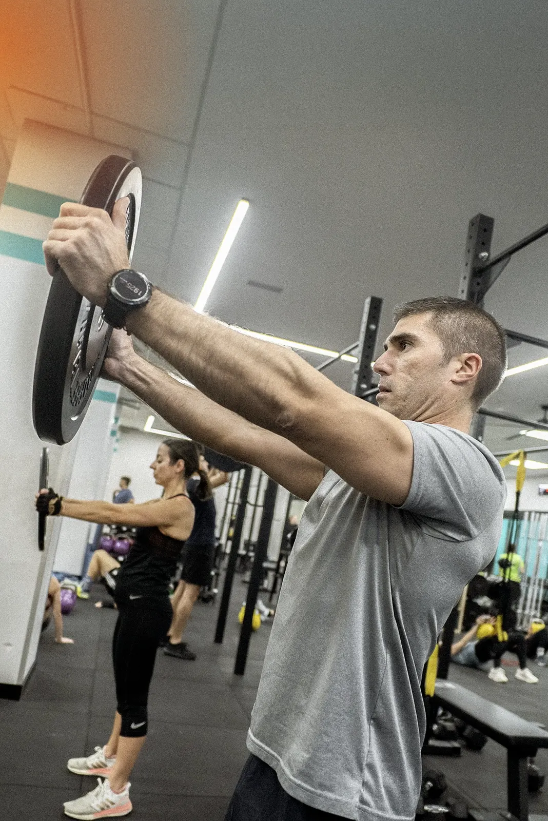 Hombre y mujer en un gimnasio sosteniendo discos de peso con los brazos estirados hacia adelante mientras realizan ejercicio en grupo.