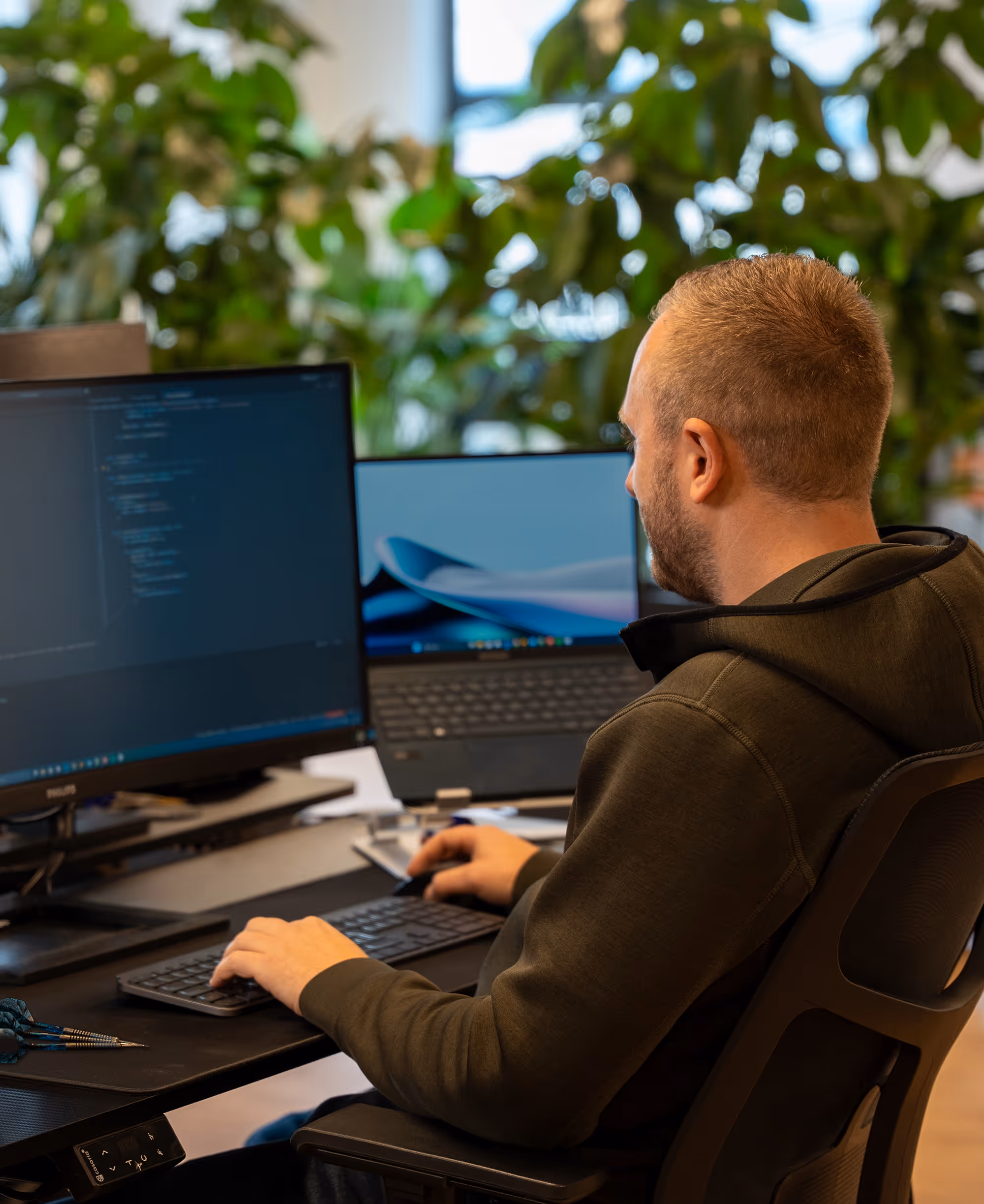 Man sitting at a desk working on a computer with dual monitors displaying code and a desktop background, surrounded by indoor plants.