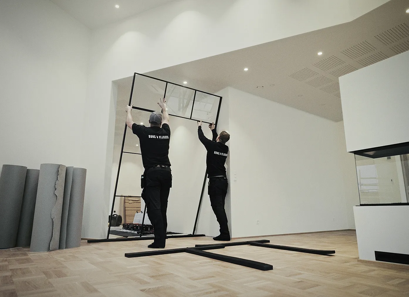 To mænd i sorte t-shirts monterer en stor rektangulær spejlramme mod en lys væg i et rum med trægulv.