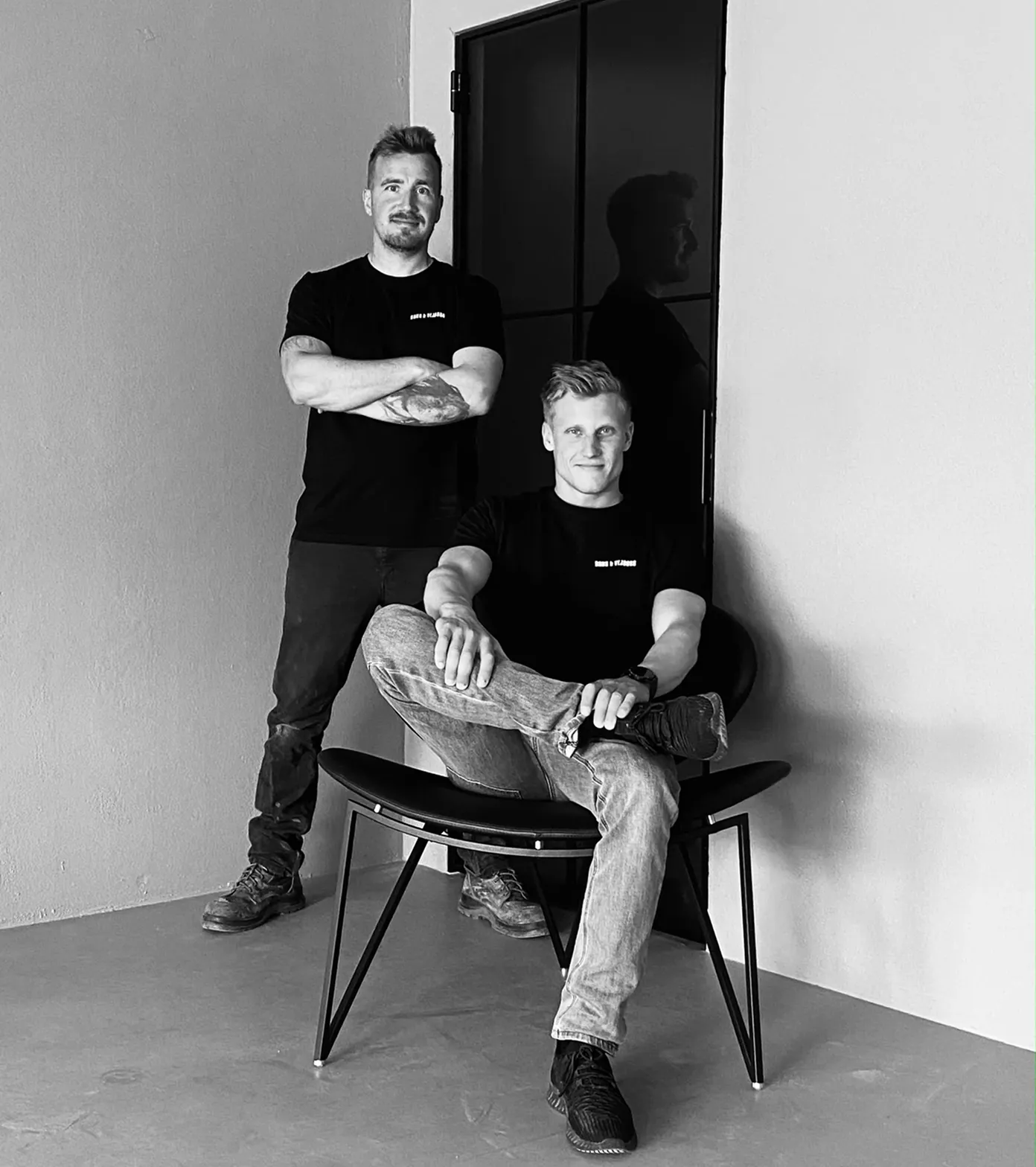 To mænd iført sorte t-shirts, den ene står med armene krydset, den anden sidder afslappet på en stol med benene krydset.