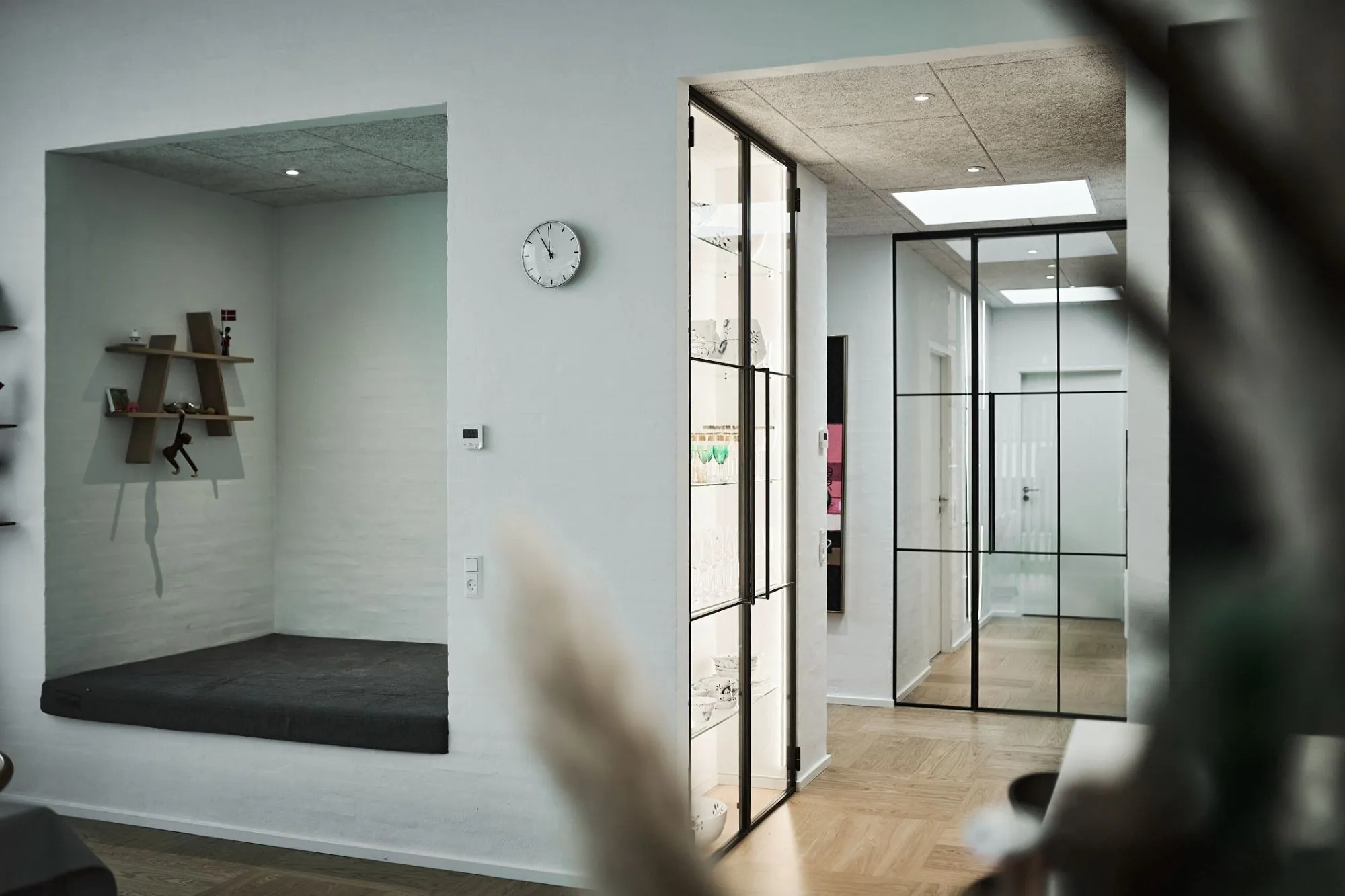 Indbygget siddeplads med sort pude og hylder med små dekorationer i et moderne, lyst rum med glasdøre og trægulv.