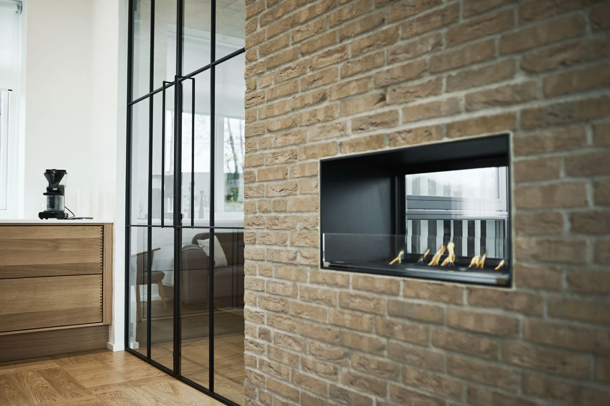 Moderne indendørs pejs med glasvindue indbygget i en murstensvæg ved siden af et køkken med træskabe og en sort kaffemaskine.