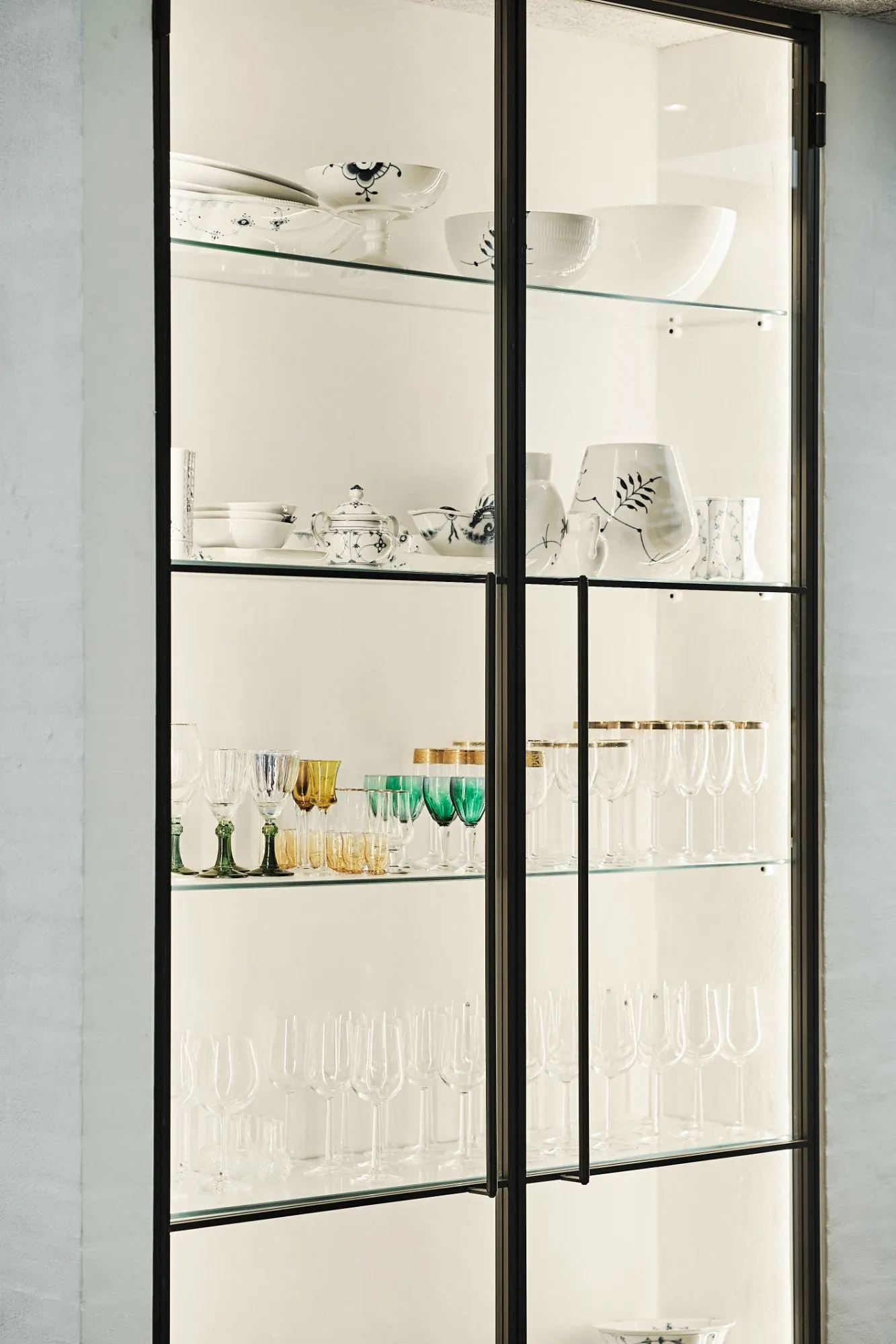 Glasvitrine med hylder, der indeholder pyntede hvide porcelænsskåle og kopper samt farvede vinglas og champagneglas med guldkant.