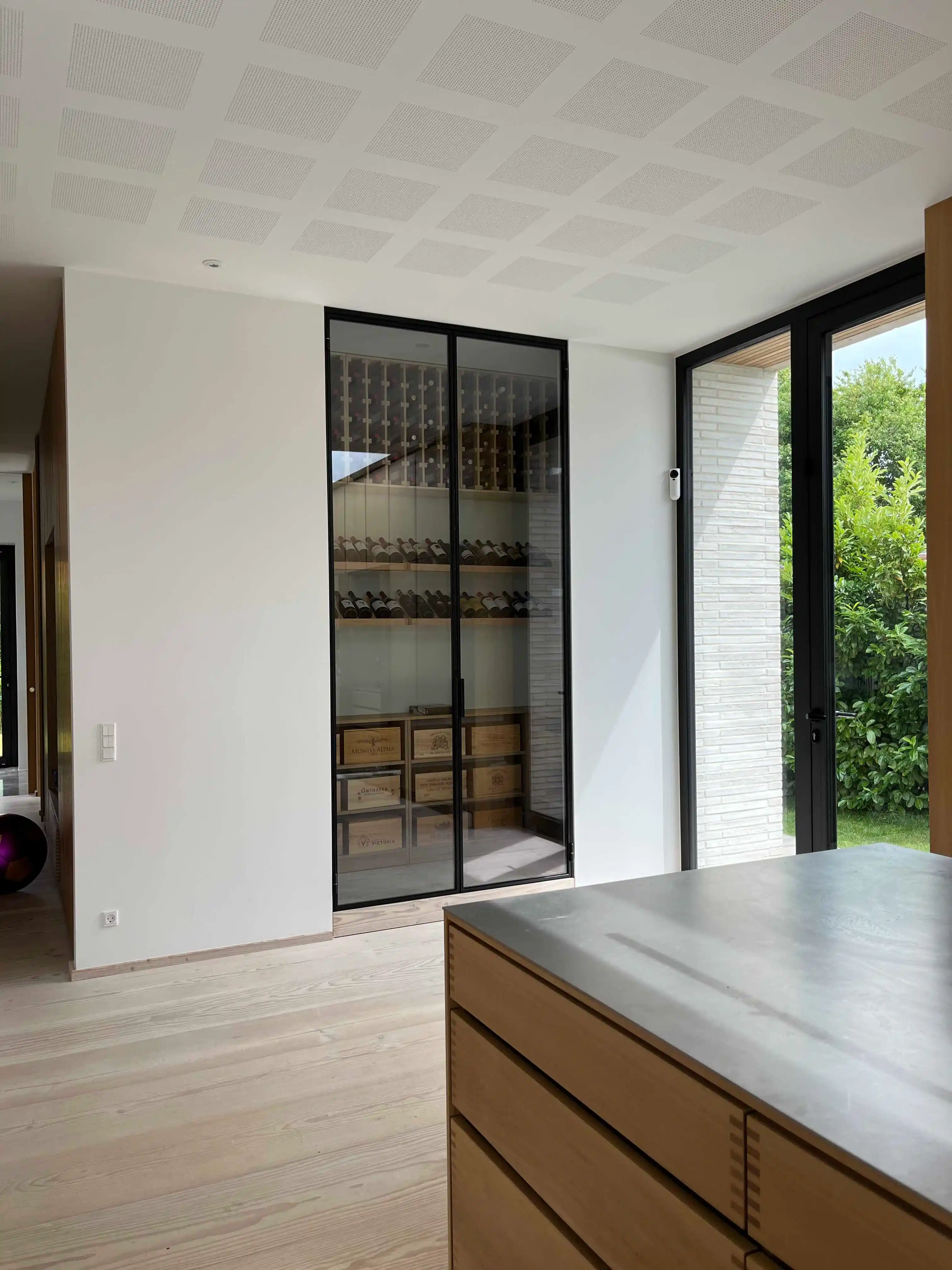 Moderne vinrum med dobbeltdør i sort metal og glas, indrettet med vinflasker og trækasser, lys trægulv og køkkenø med betonbordplade.