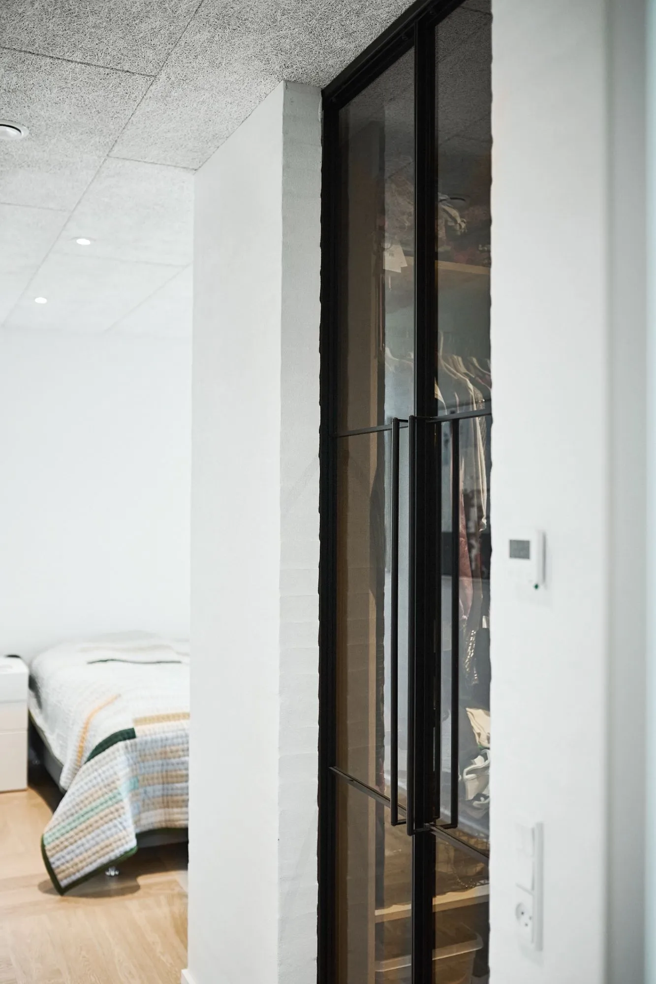 Garderob med sorte glasdøre ved siden af et hvidt soveværelse med en seng dækket af et stribet tæppe.