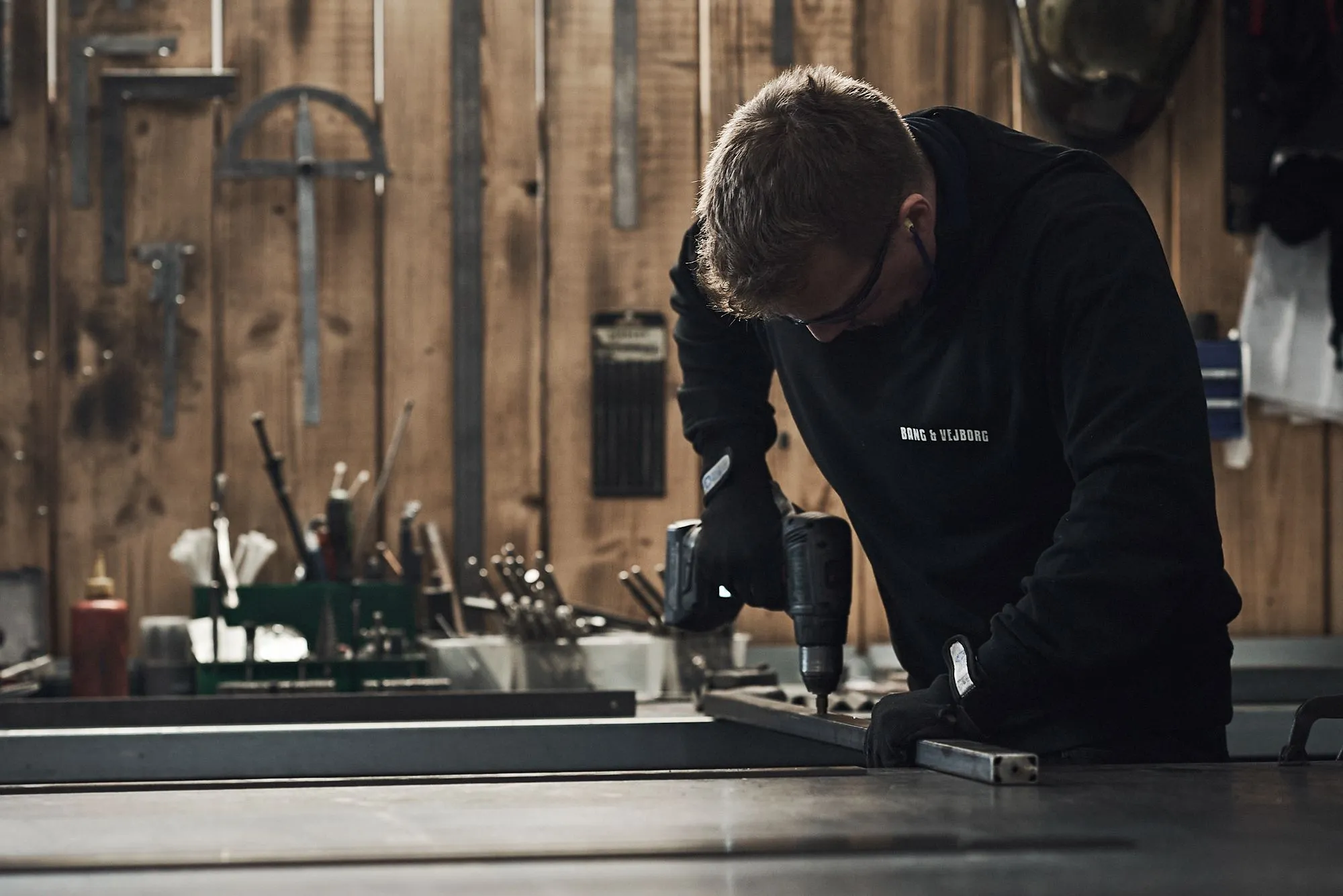 Mand med beskyttelsesbriller bruger en elektrisk boremaskine på et metalstykke i et værksted.