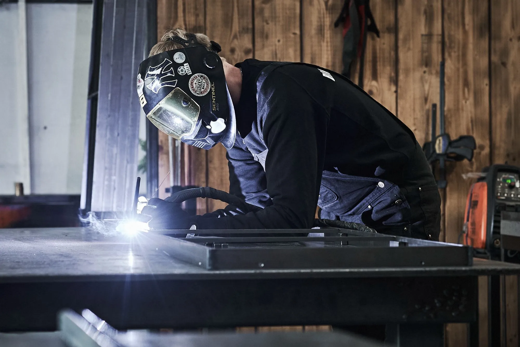 Svejser iført beskyttelsesmaske og handsker arbejder med metal i en værkstedsmiljø.