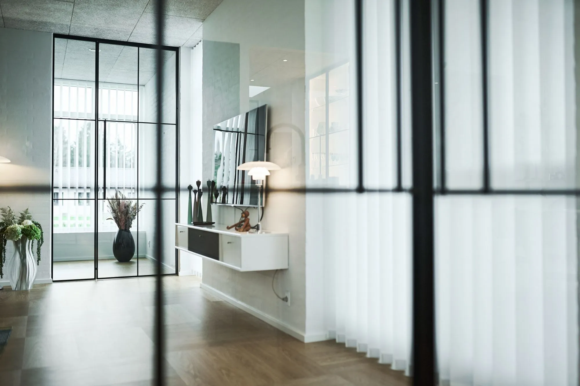 Moderne stue med gulv-til-loft vinduer med hvide lamelgardiner, hvid væghængt skænk, dekorative vaser og skulpturer, og trægulv.