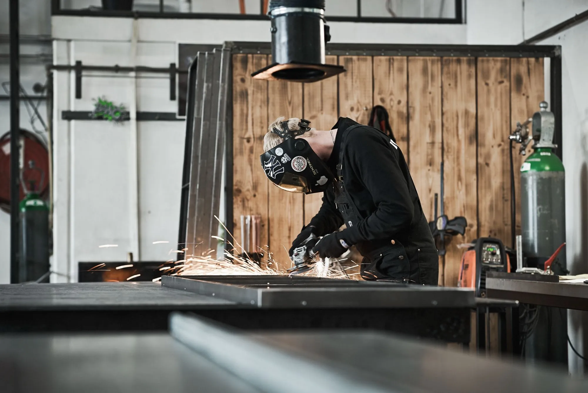 Person iført svejsehjelm og handsker, der arbejder med vinkelsliber, hvilket skaber gnister i et værksted.