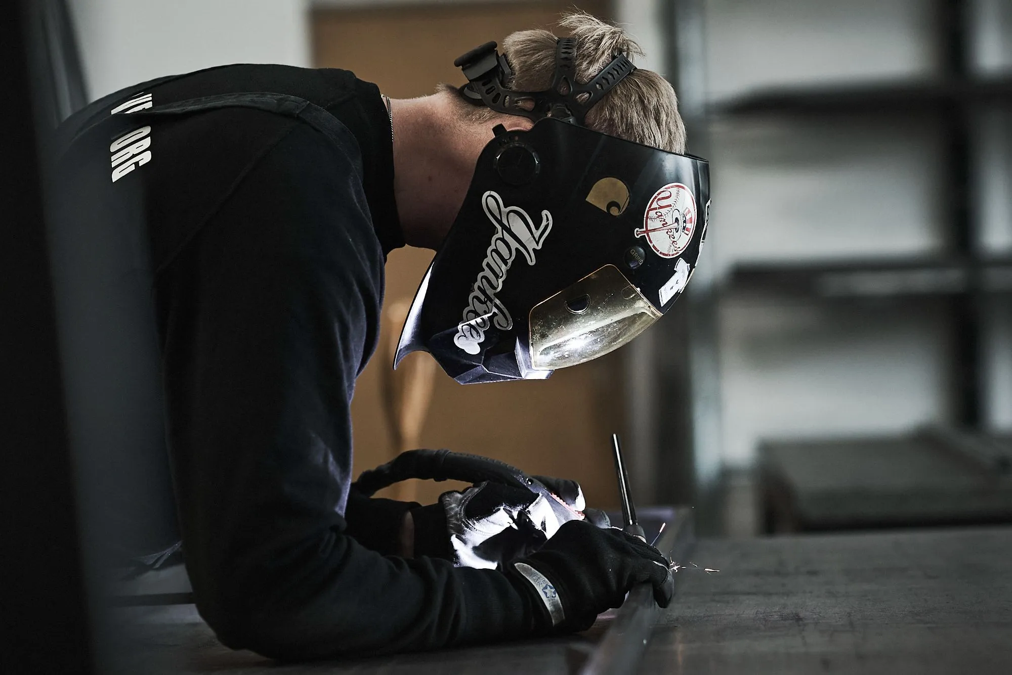 Person iført svejsehjelm og handsker svejser metal i en industribygning.