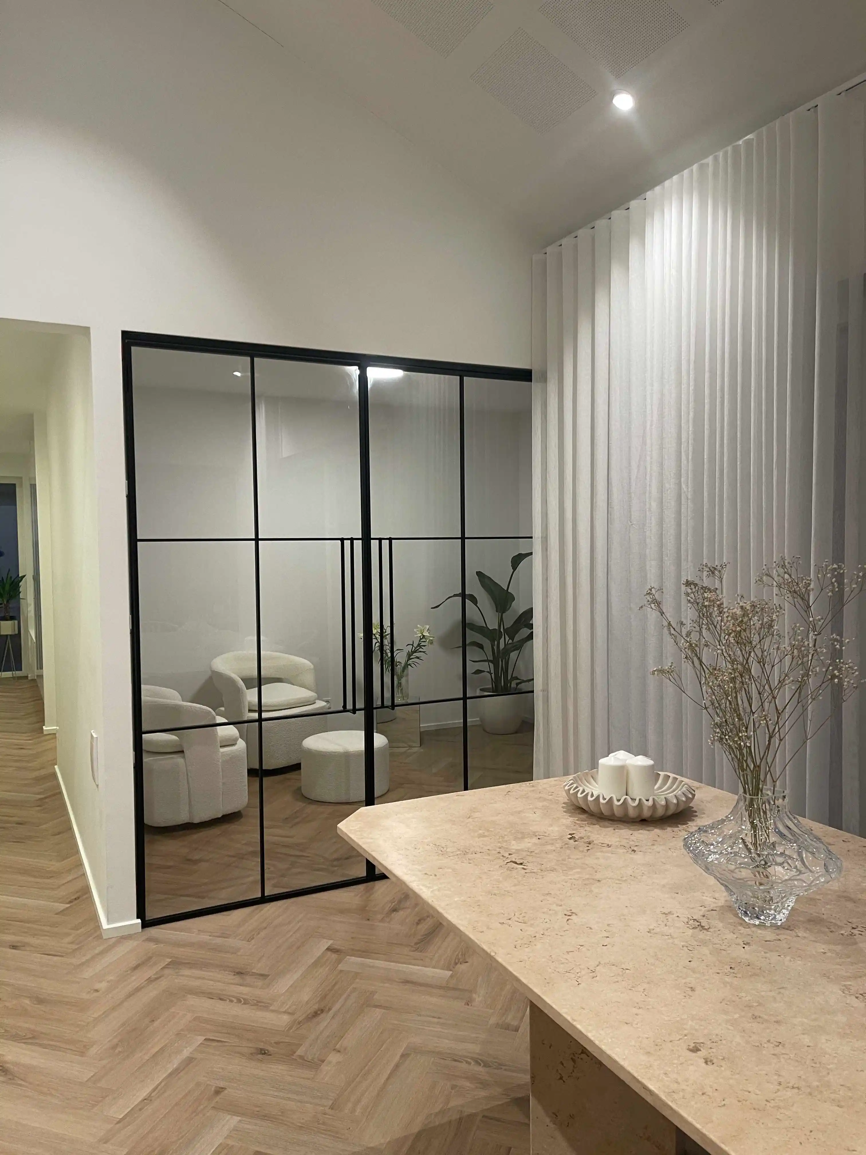 Moderne indretning med sorte dobbeltdøre i glas, to beige polstrede lænestole, et rundt puf, og plante i hvid potte, forbundet med borde i beige sten med lysestager og en klar vase med tørrede blomster.