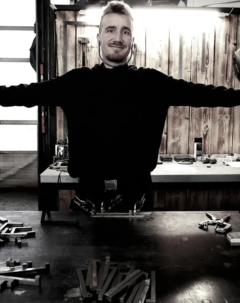 Ung mand med hænderne udstrakte iført mørkt tøj, stående i en træværksted med værktøj og materialer på bordet foran sig.