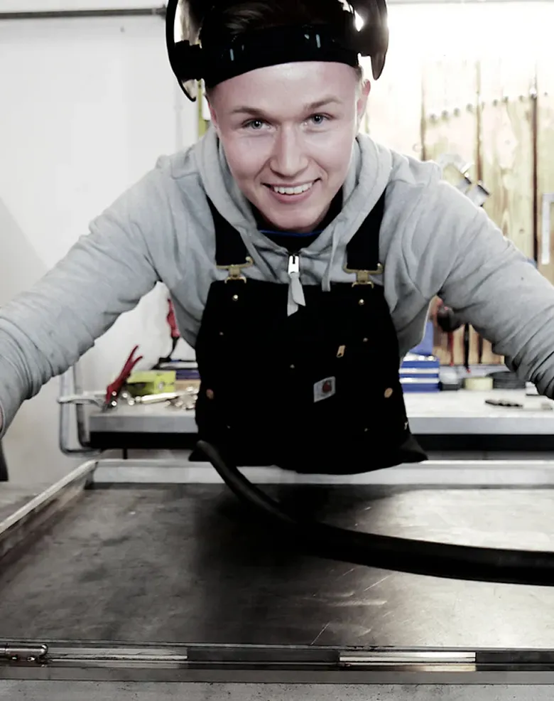 Ung mand i arbejdstøj smilende over en arbejdsbænk med værktøj i baggrunden.