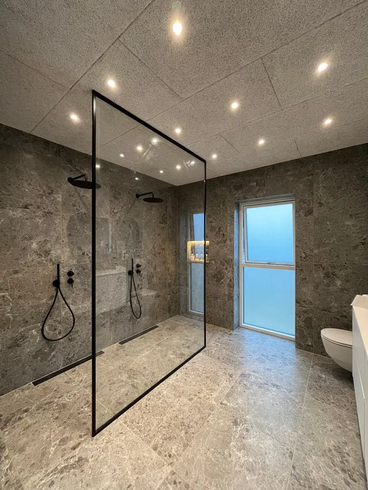 Moderne badeværelse med grå marmorgulv og vægge, glas brusevæg med sort ramme og sort brusehoved, stor frostet vindue og toilet.