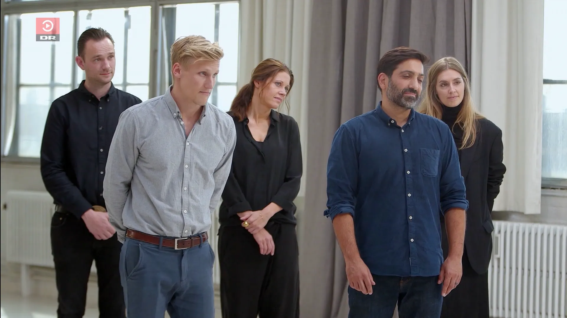 Fem voksne mennesker i afslappet tøj står i et lyst rum med store vinduer og grå gardiner.