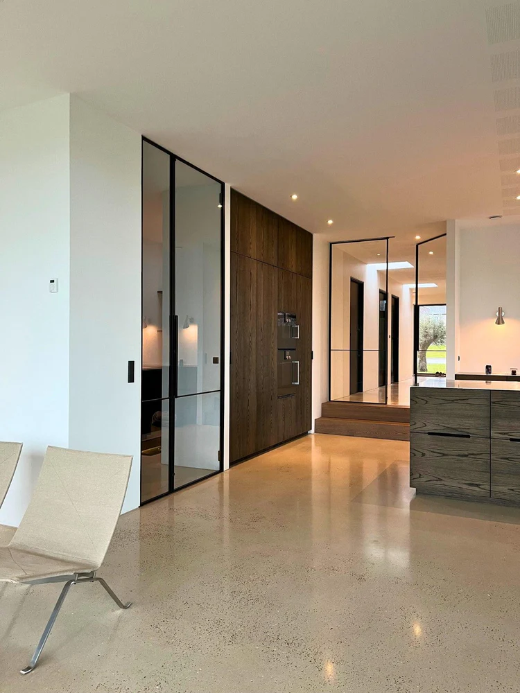 Moderne indgang med glasdør, mørkt træpanel, trin op til gang og beige stole i forgrunden.