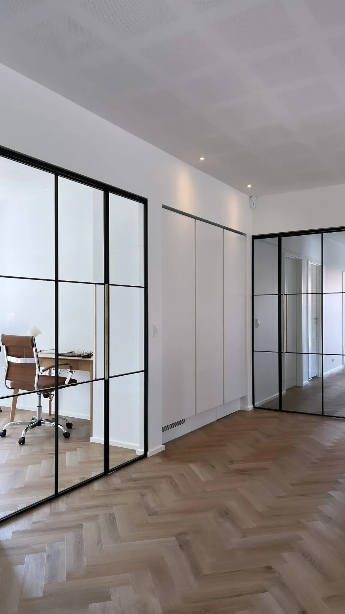 Moderne kontor med glasvægge i sort ramme, brunt kontorstol og skrivebord med lamper, samt lyst trægulv i sildebensmønster.