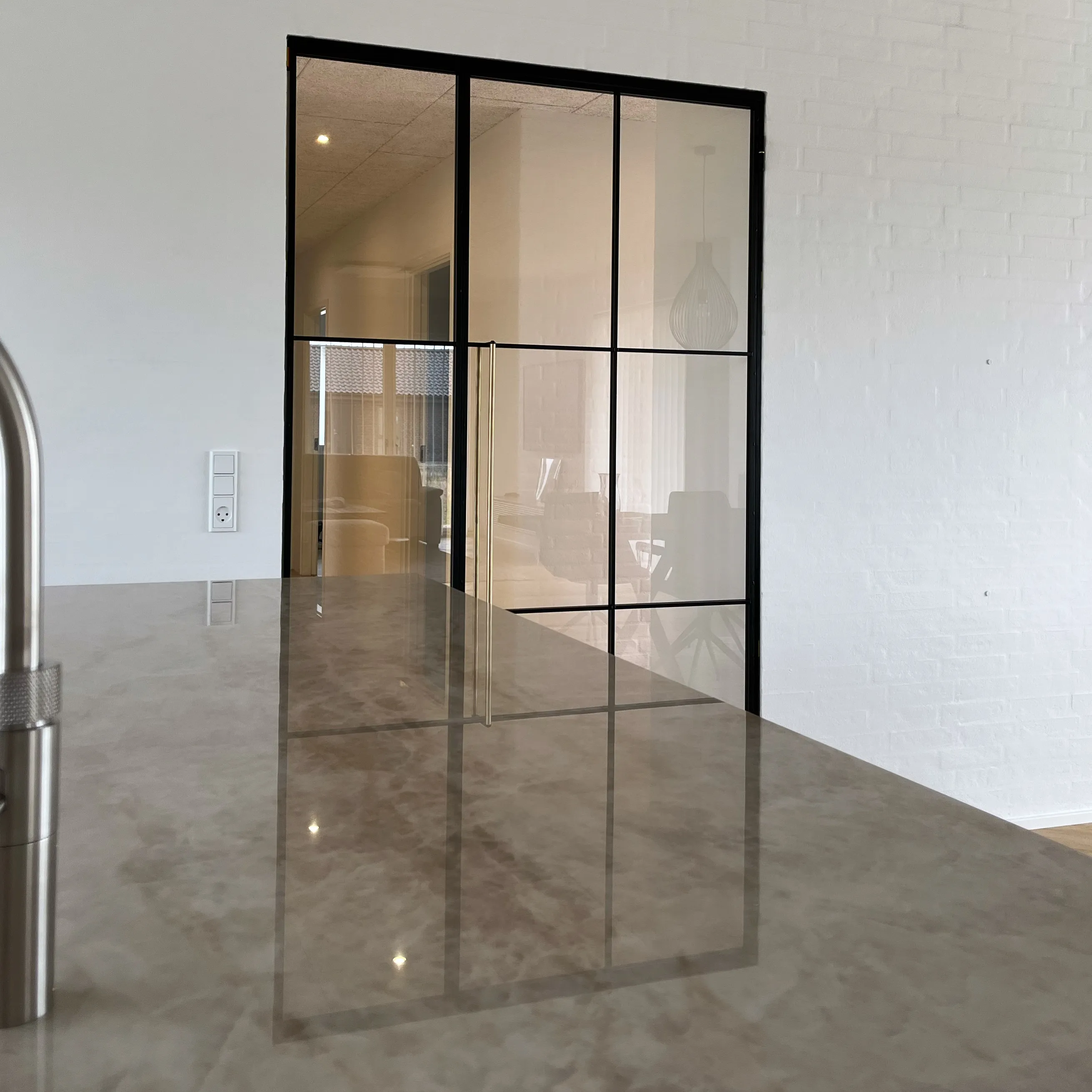 Et moderne køkkenbord i marmor med vandhane og sort metalramme glasdør i baggrunden, der viser en stue.