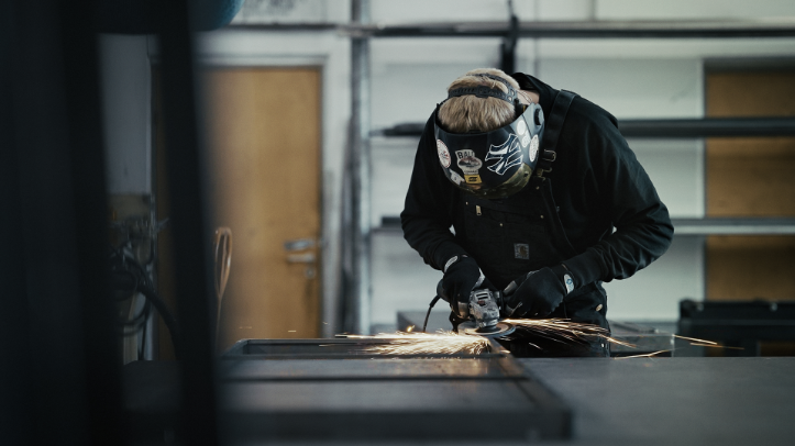 En person iført svejsehjelm og handsker sliber metal, mens gnister flyver i et værksted.