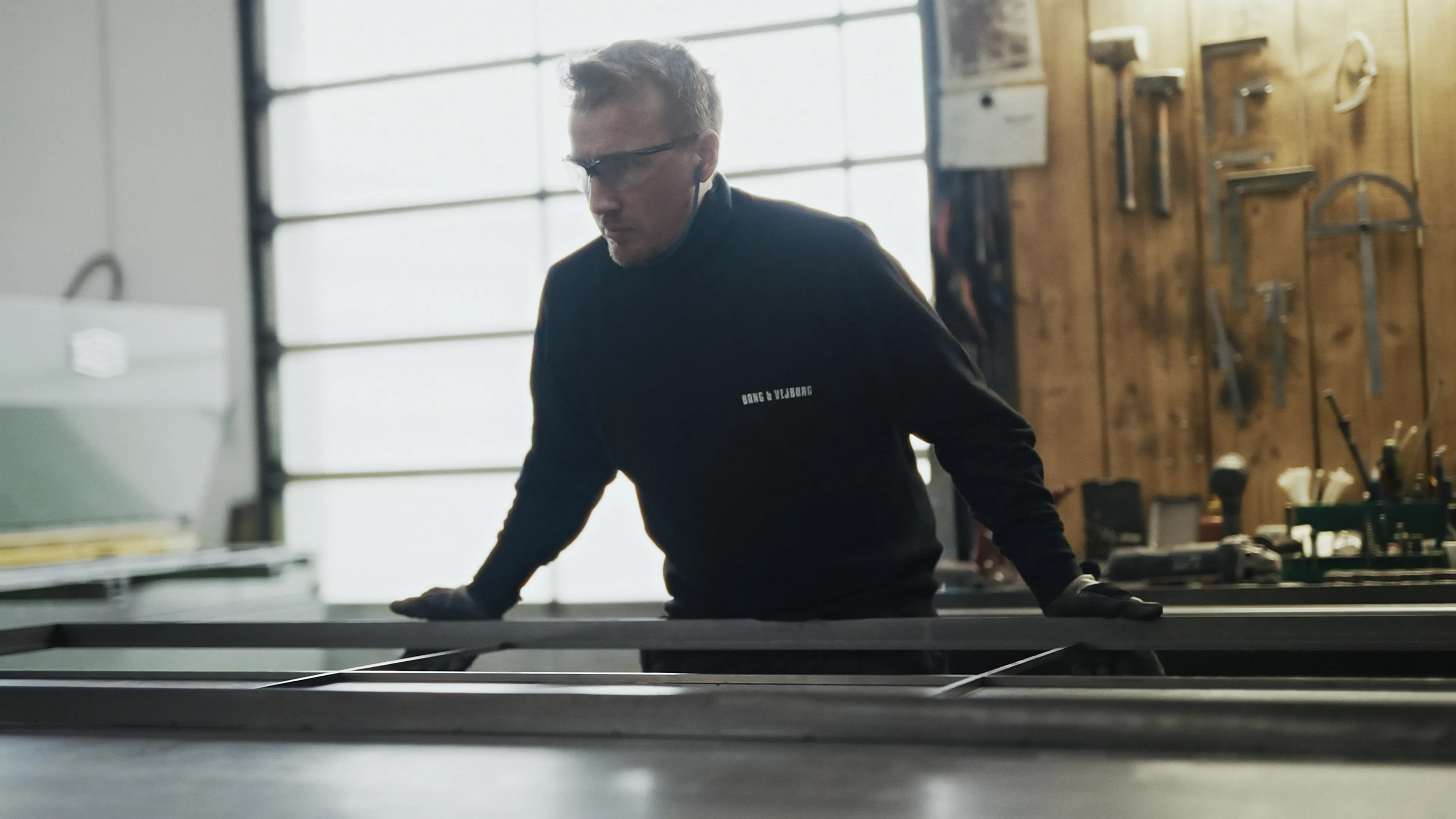 Mand med beskyttelsesbriller og handsker arbejder med metalramme i værksted.