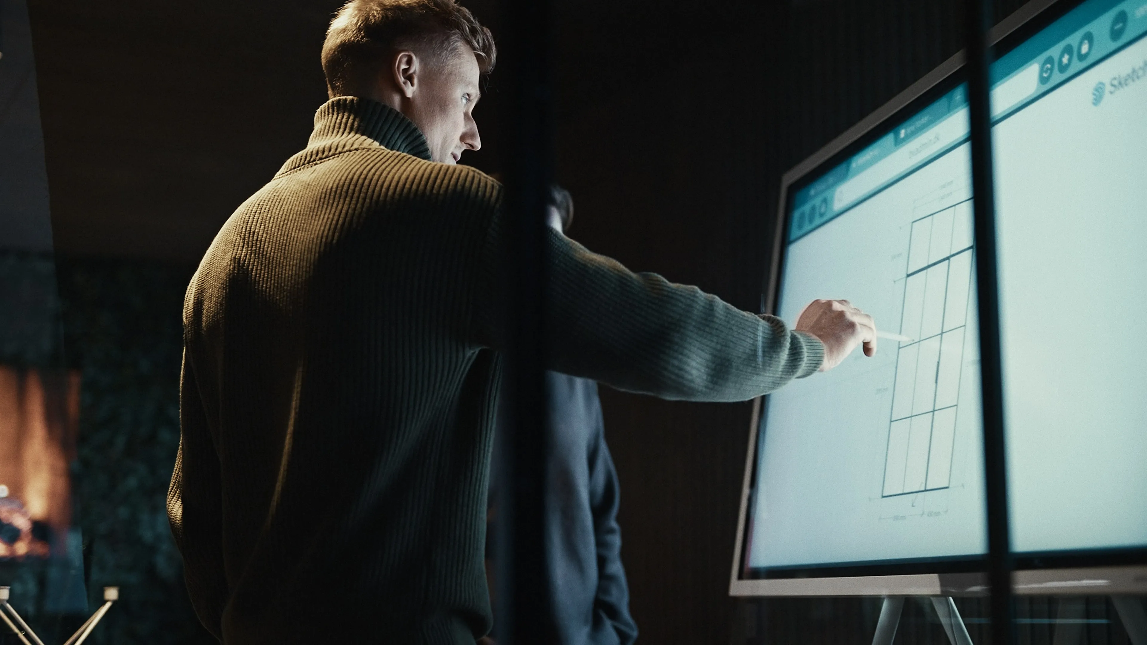 Mand i grøn ribstrikket trøje peger på diagram på stor skærm med en stylus.