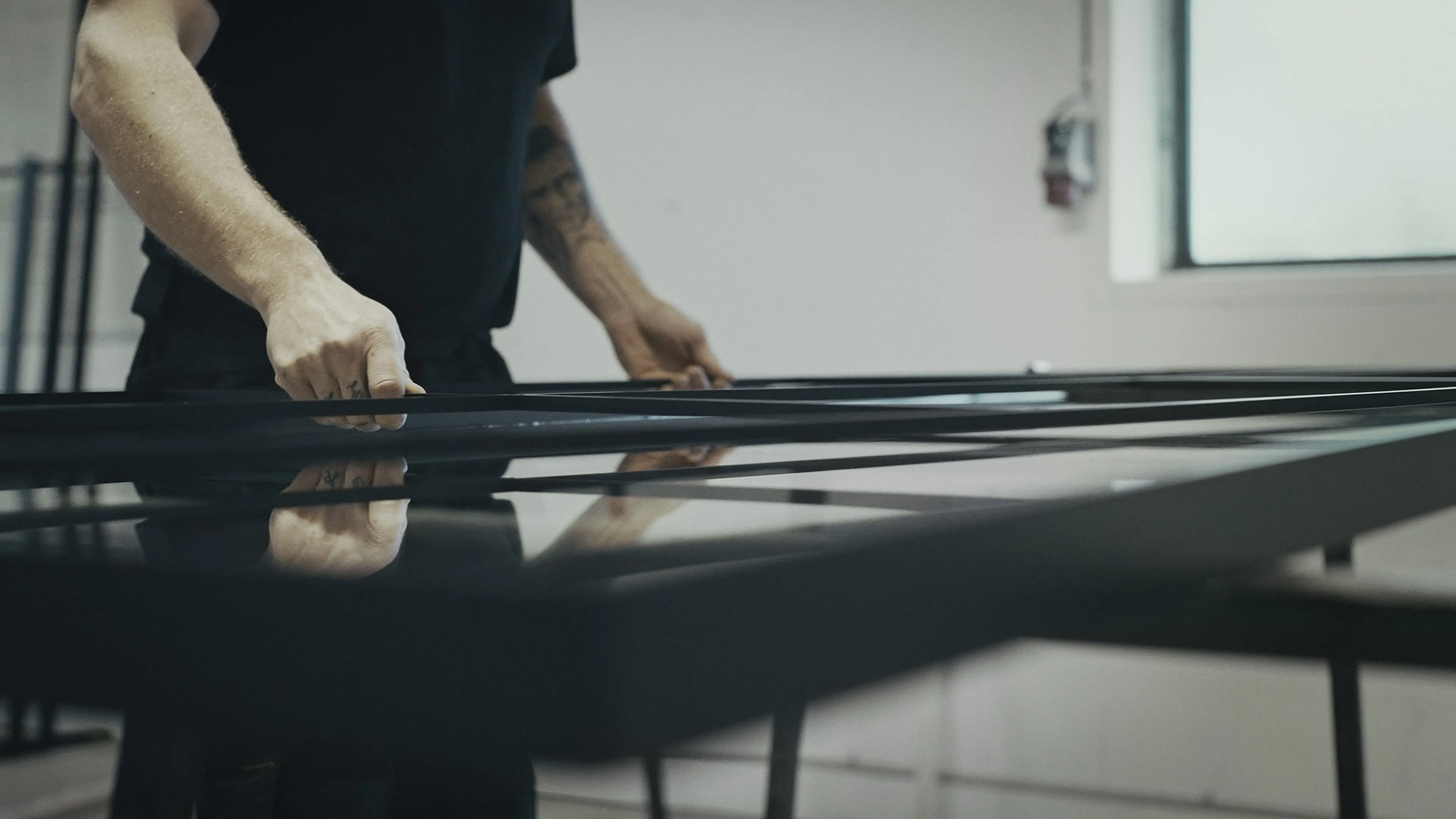 Person iført sort t-shirt, der håndterer eller monterer sorte metal- eller glasrammer på et bord indendørs.