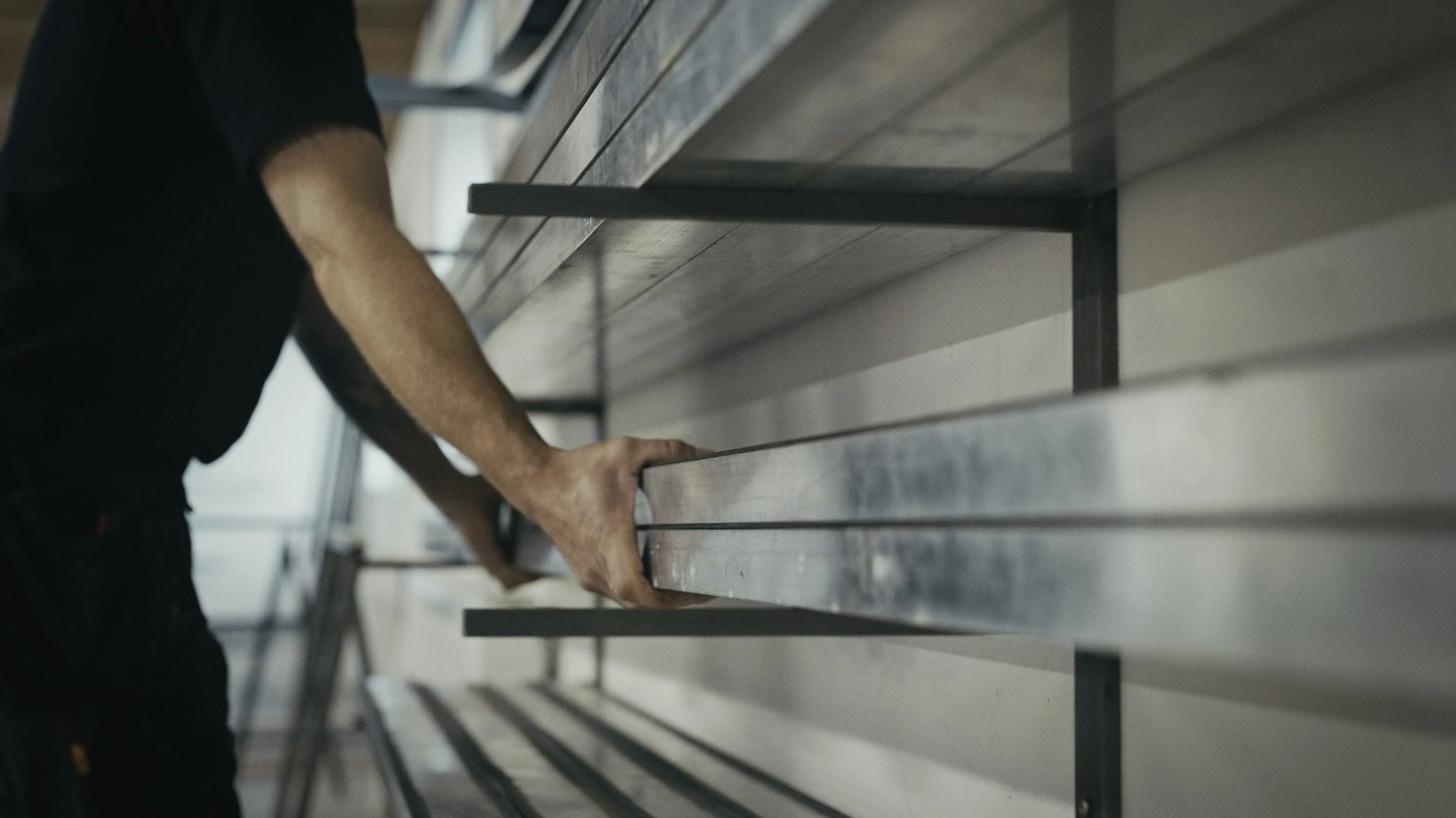 En person med sort t-shirt placerer metalstænger på hylder i et rum med naturligt lys.