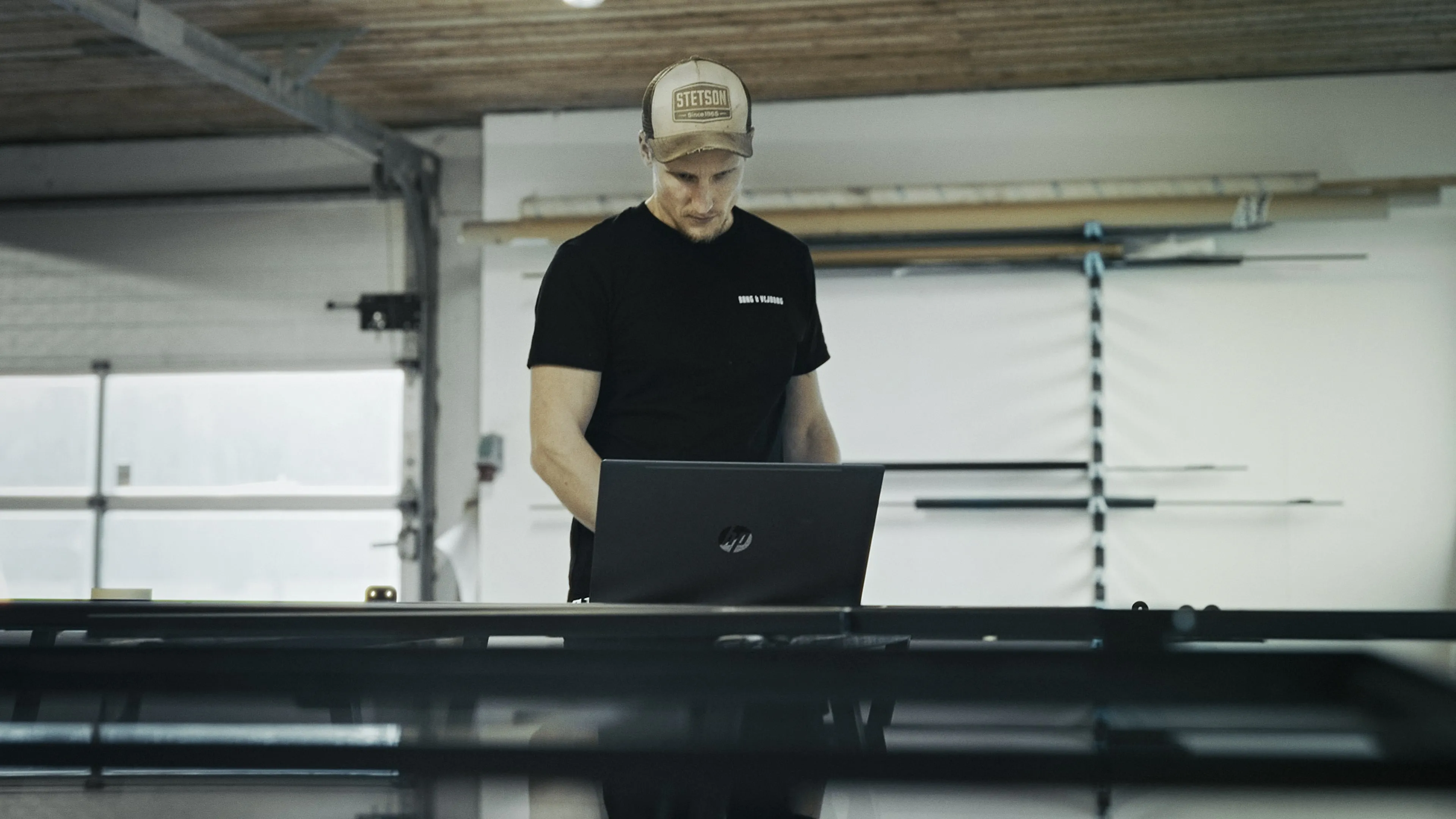 Mand i sort t-shirt og kasket arbejder koncentreret på en HP bærbar i et lyst værksted.