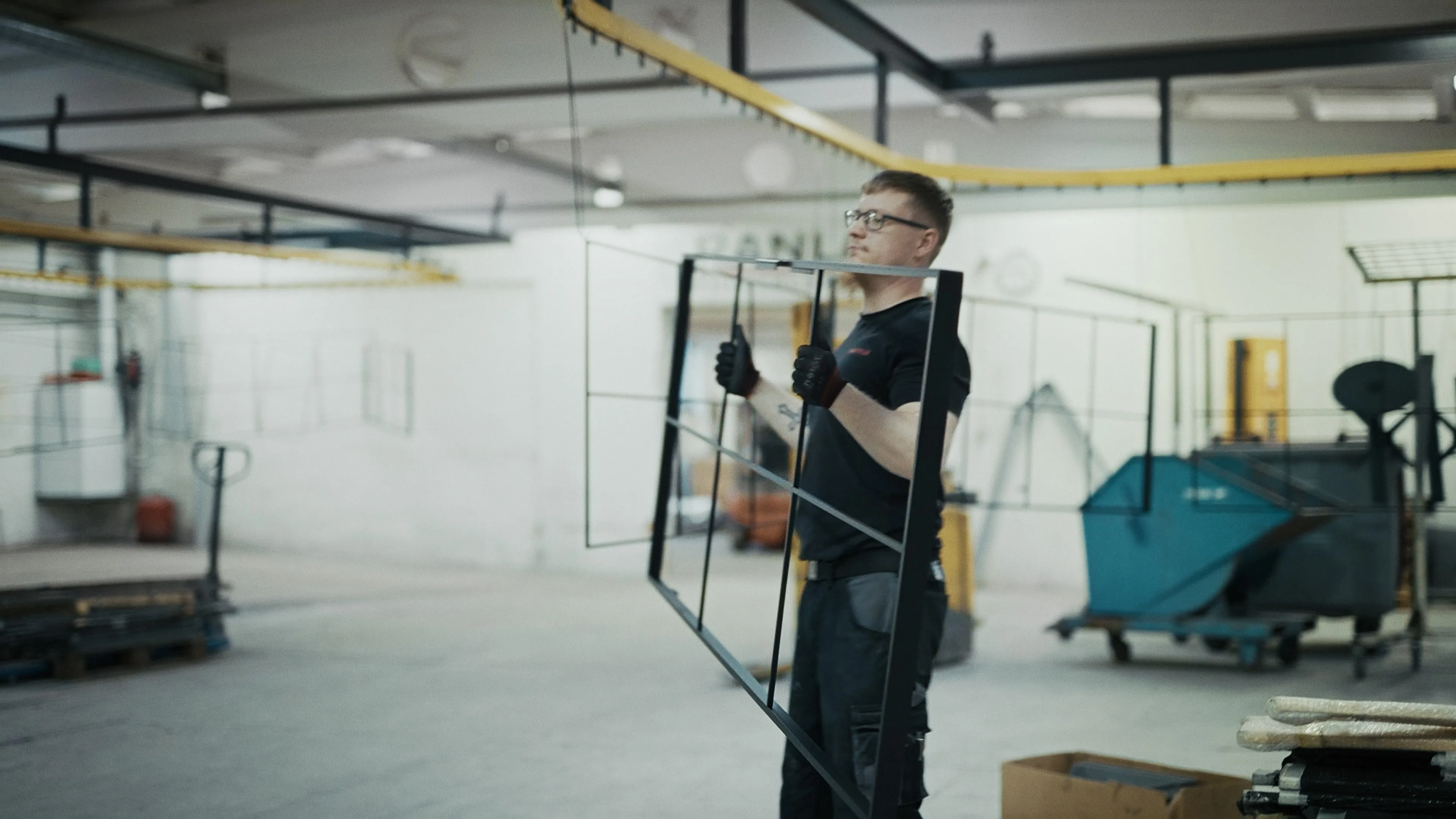 Mand iført sikkerhedshandsker bærer en stor metalramme i en industribygning.