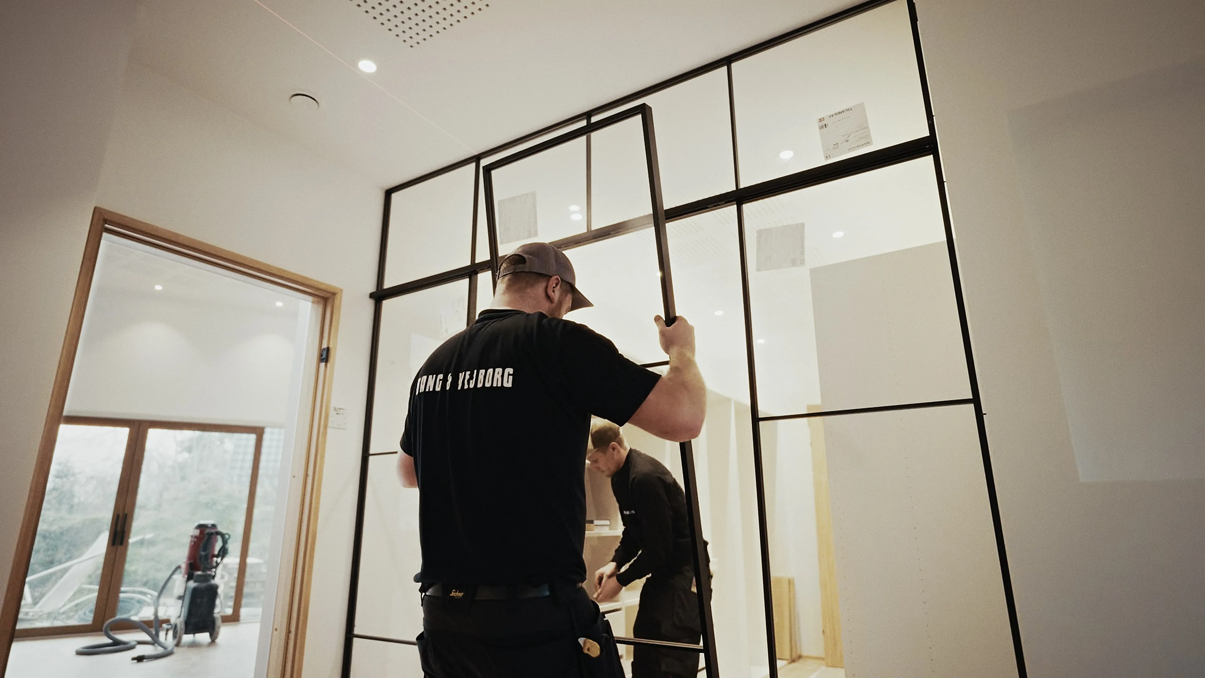 To håndværkere i et lyst rum, hvor den ene holder en sort metalskillevæg, mens den anden arbejder med et møbel i baggrunden.