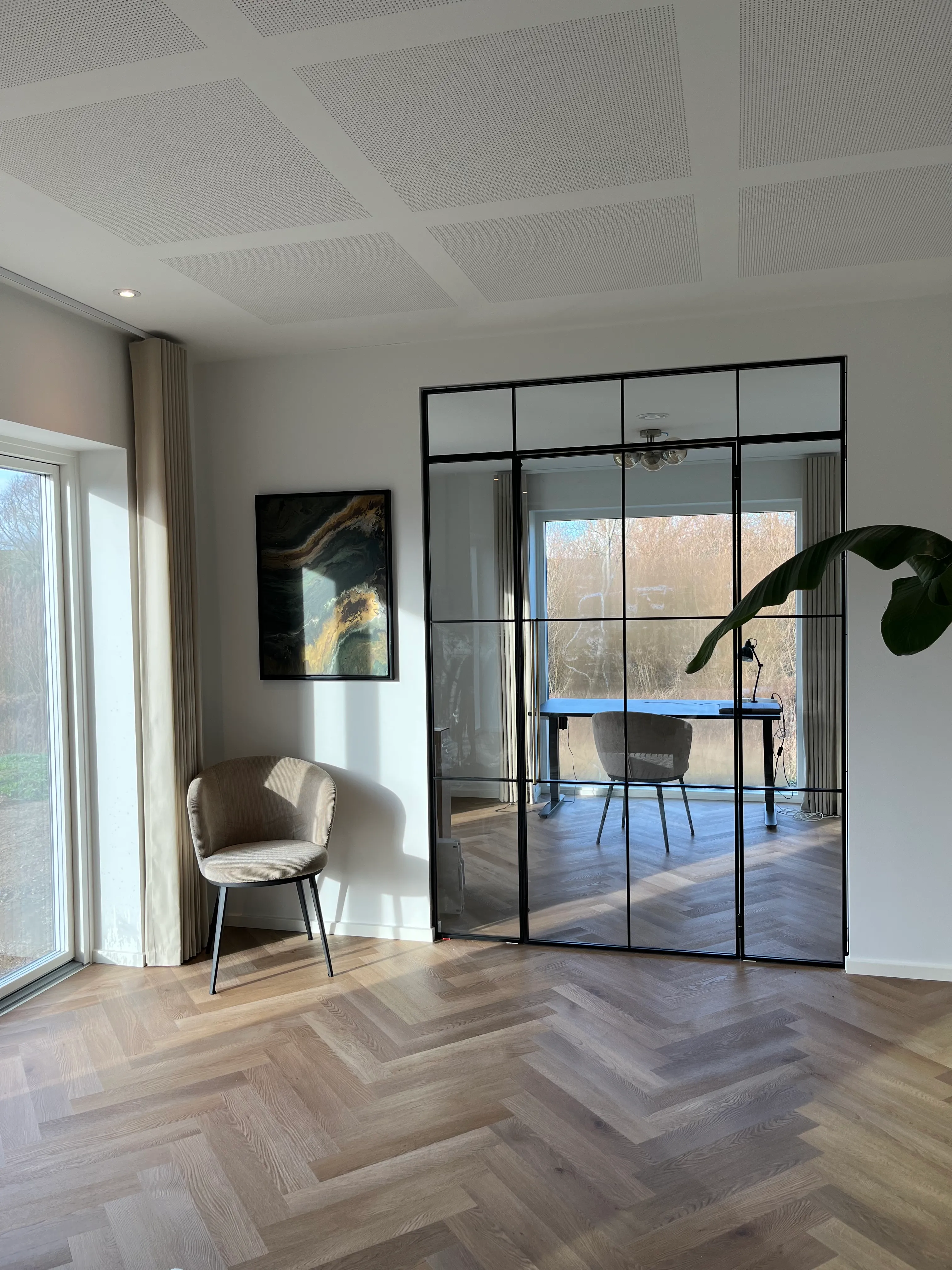 Lys stue med beige stol ved vindue og glasvæg, der reflekterer et skrivebord med stol foran et stort vindue.