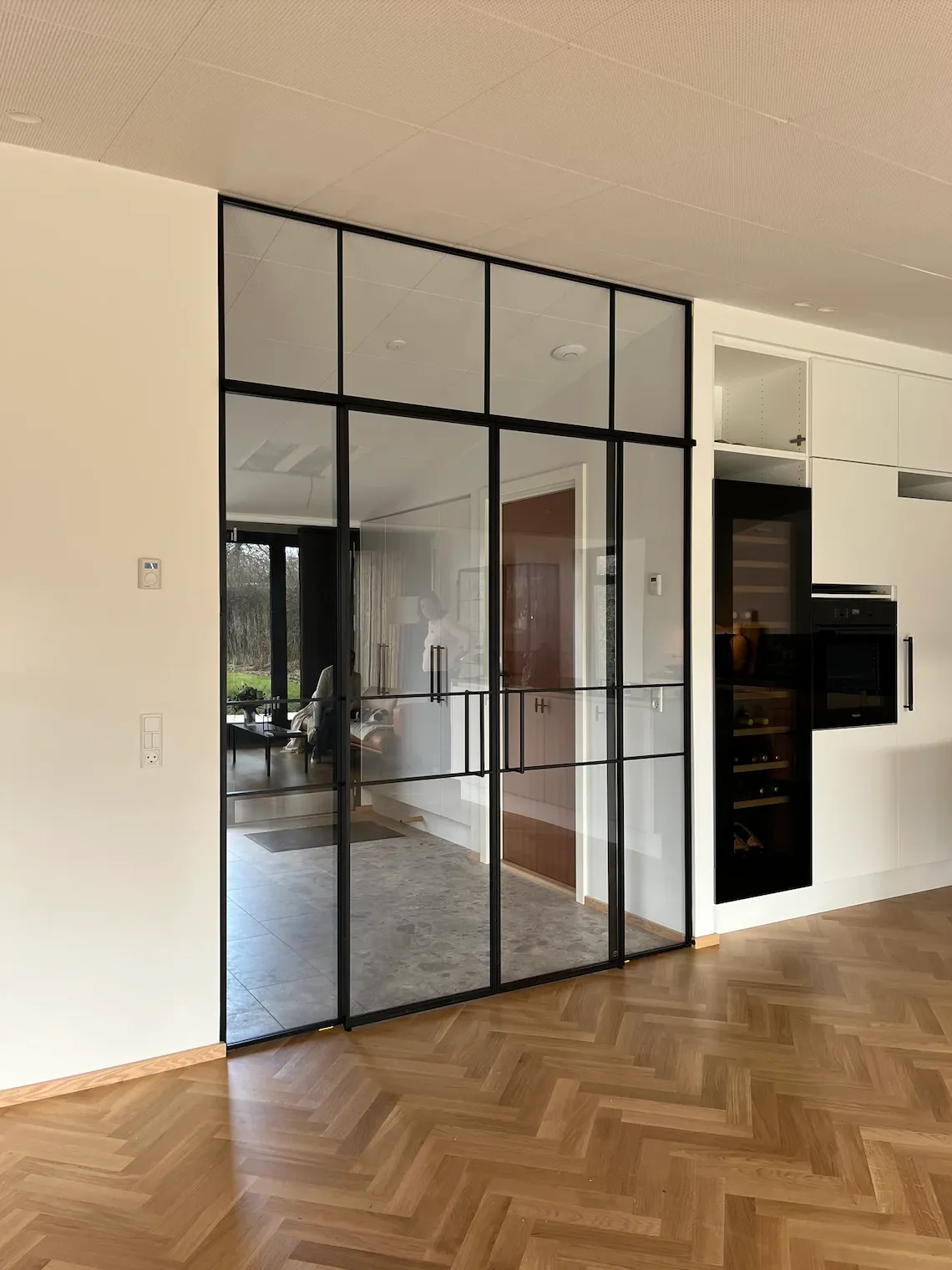 Moderne hvide køkkenkabe med integreret ovn og vinkøleskab, adskilt fra en stue med glasvæg og sorte metalrammer, med lyst trægulv i forgrunden.