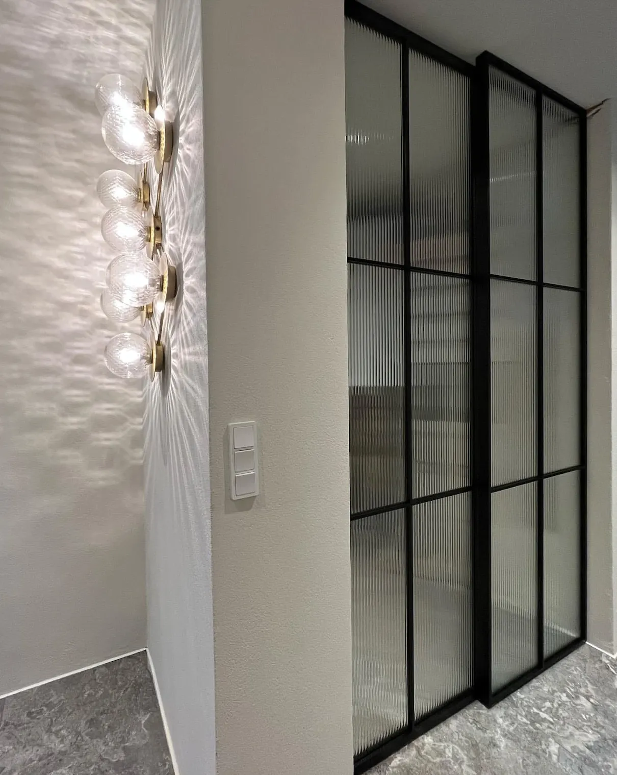 Moderne væglampe med seks lampeglas, der kaster dekorative skygger, ved siden af en sort ramme med ribbet glasdør i et rum med marmorgulv.